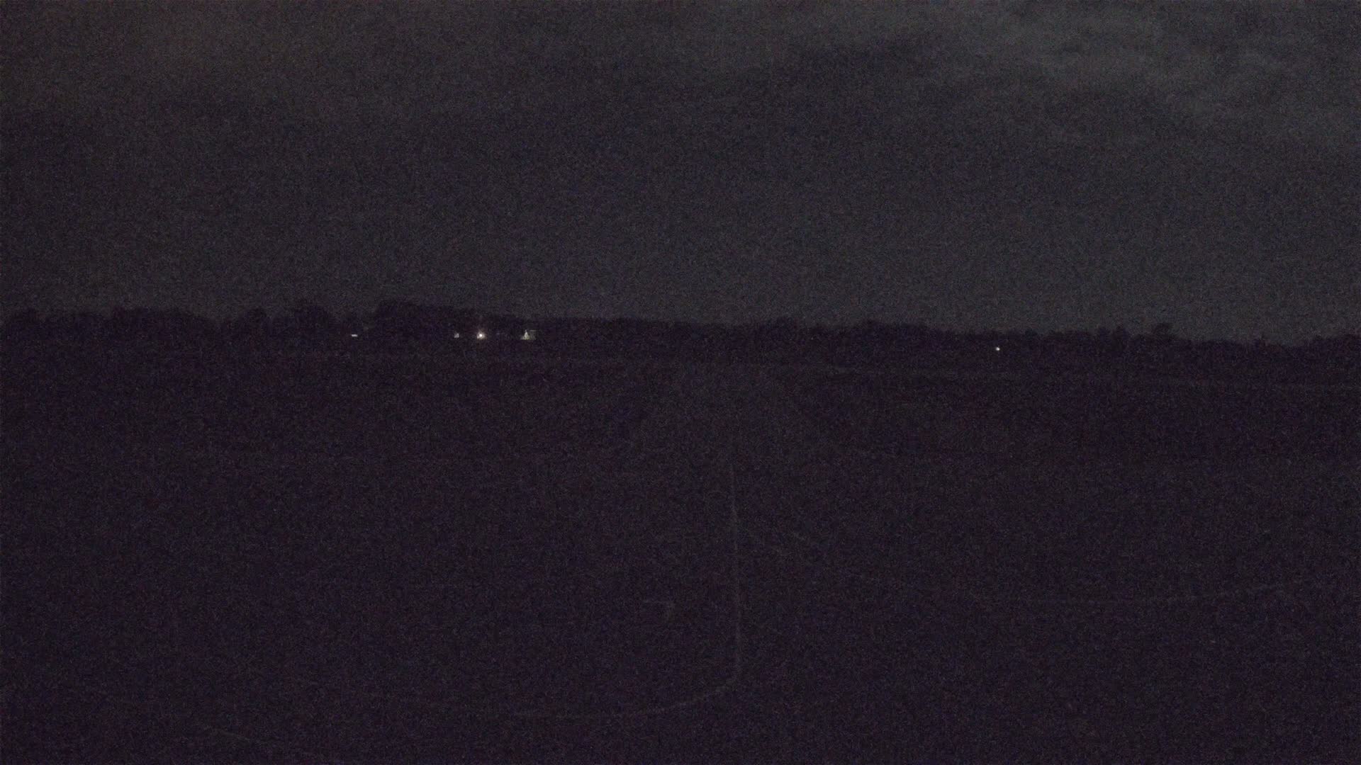 A dark, nighttime landscape shows a distant line of trees and a few faint lights under a cloudy sky.
