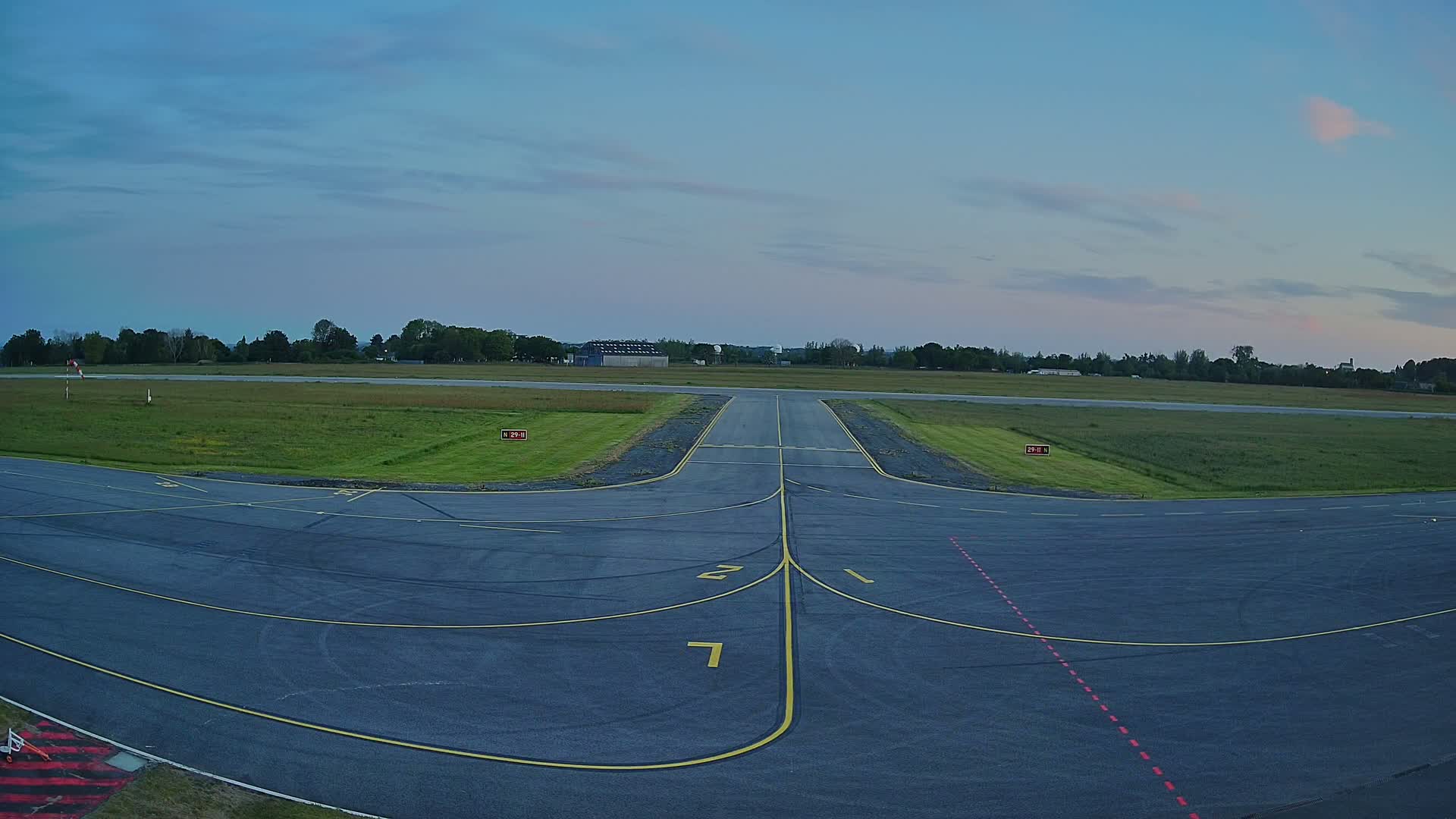 An airfield's tarmac, marked with yellow lines, leads to a grassy runway under a clear, twilight sky.