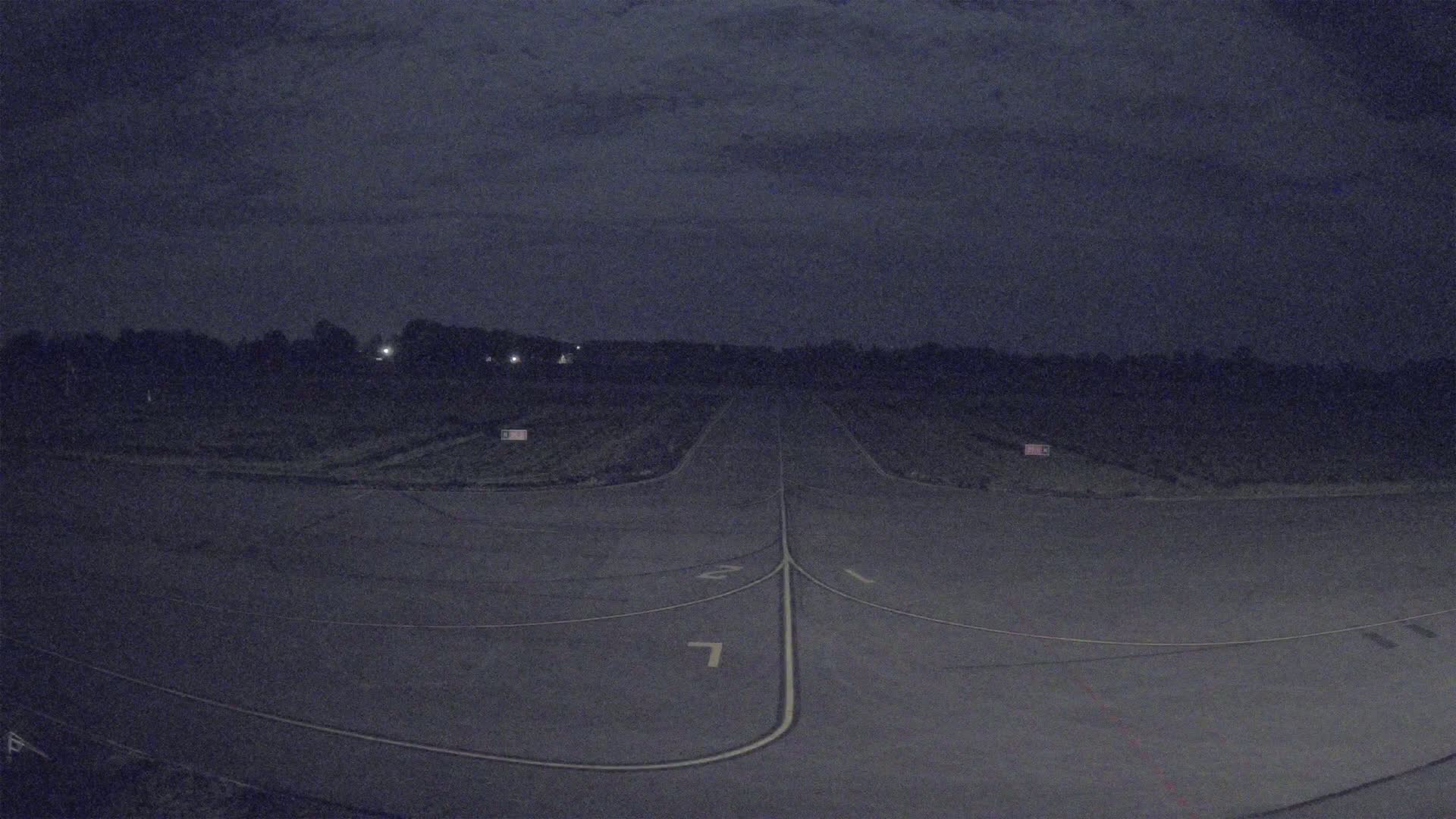 A dark asphalt runway extends into a treeline under a mostly cloudy night sky.