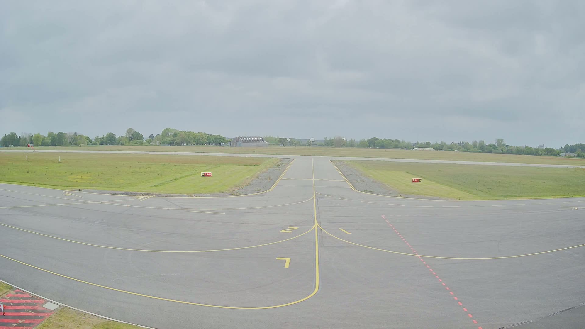A paved airport runway and taxiway intersect under an overcast sky, surrounded by grassy fields and trees.