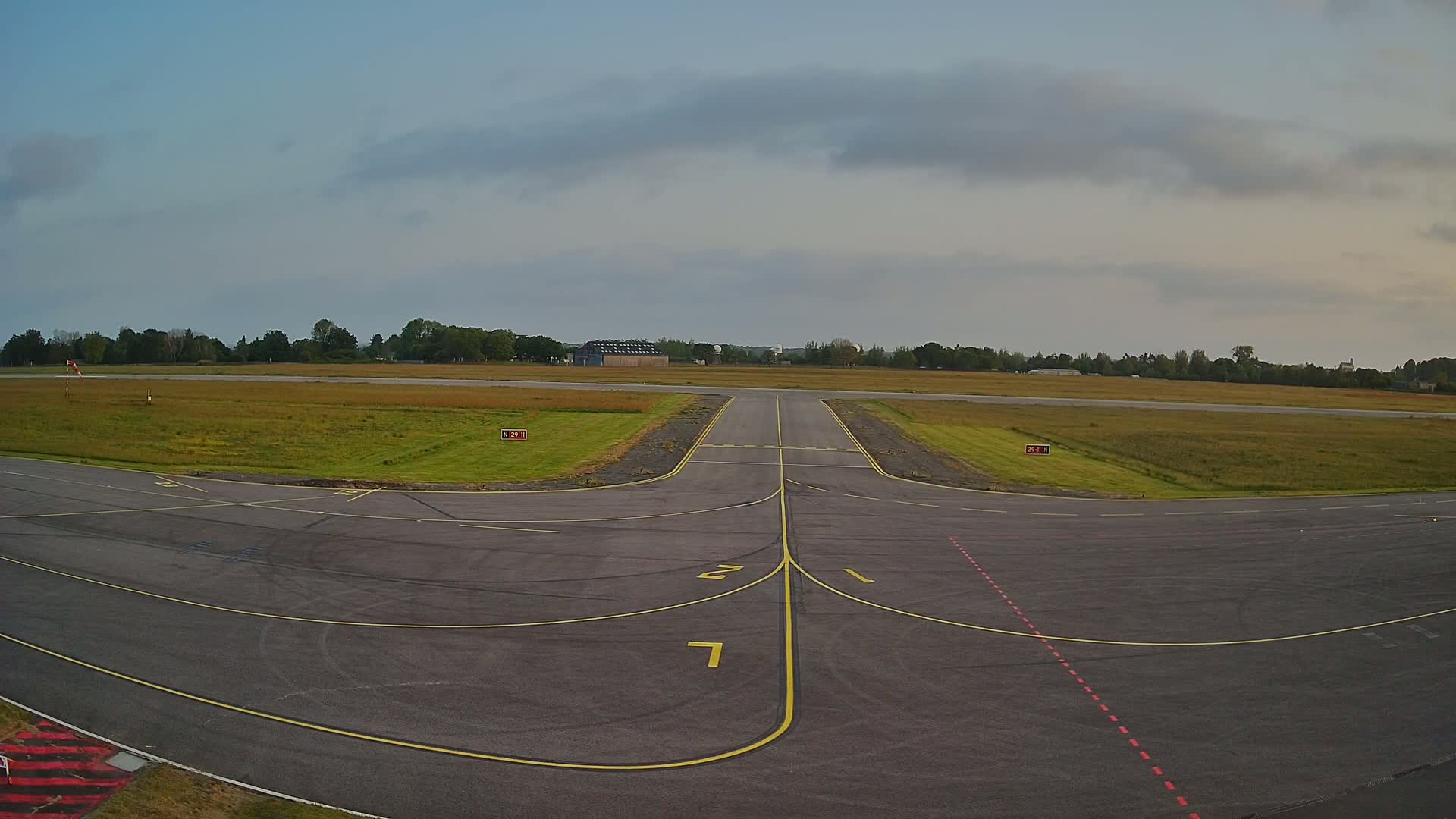 A mostly clear sky overlooks a tarmac runway intersection at an airfield, with grassy fields surrounding the paved area.