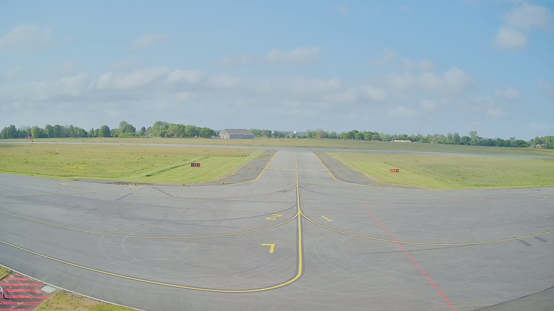 An empty airport tarmac with yellow lines and a runway extending to a grassy field under a partly cloudy sky.