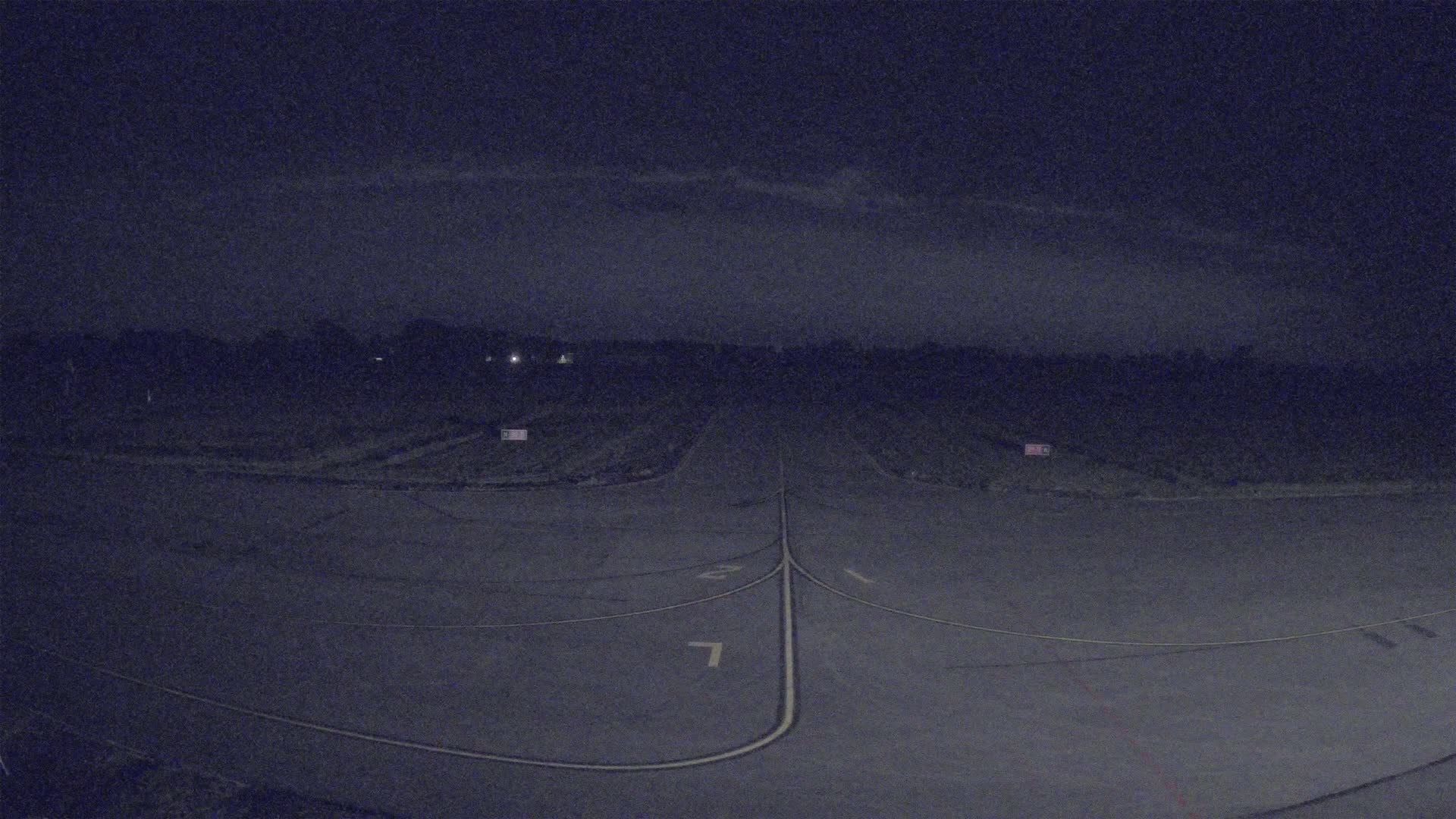 A nighttime aerial view of a paved runway, bordered by dark vegetation under a mostly clear, dark sky.