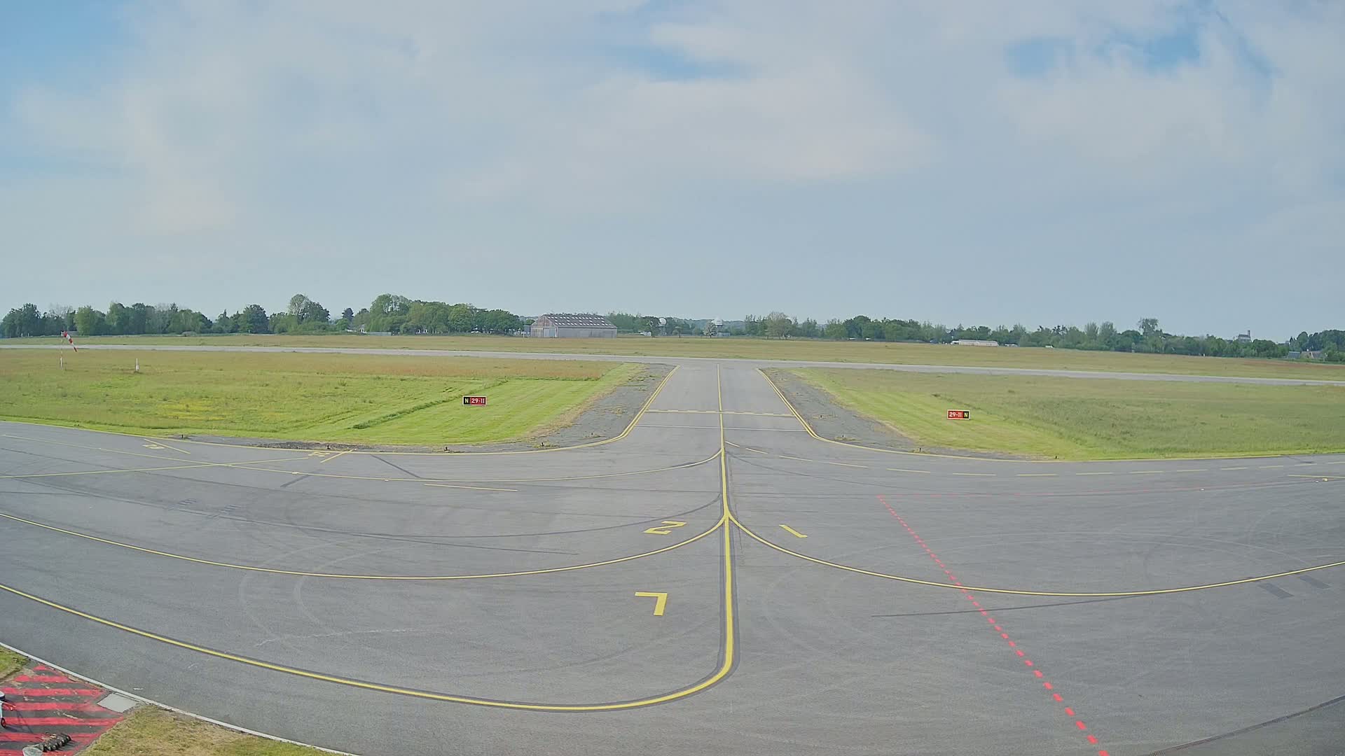 A paved airport taxiway with yellow lines intersects a grassy runway under a partly cloudy sky.
