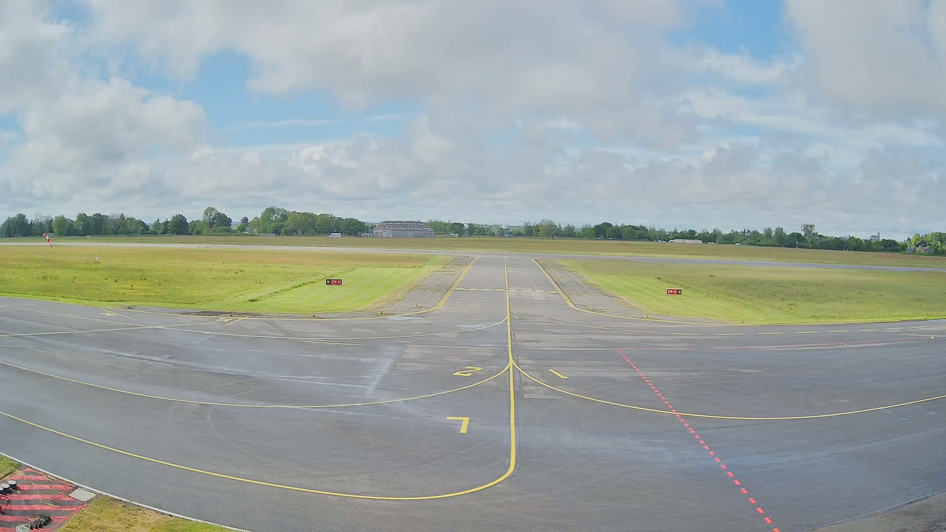 An airport runway with a grassy field and trees in the background under a partly cloudy sky.
