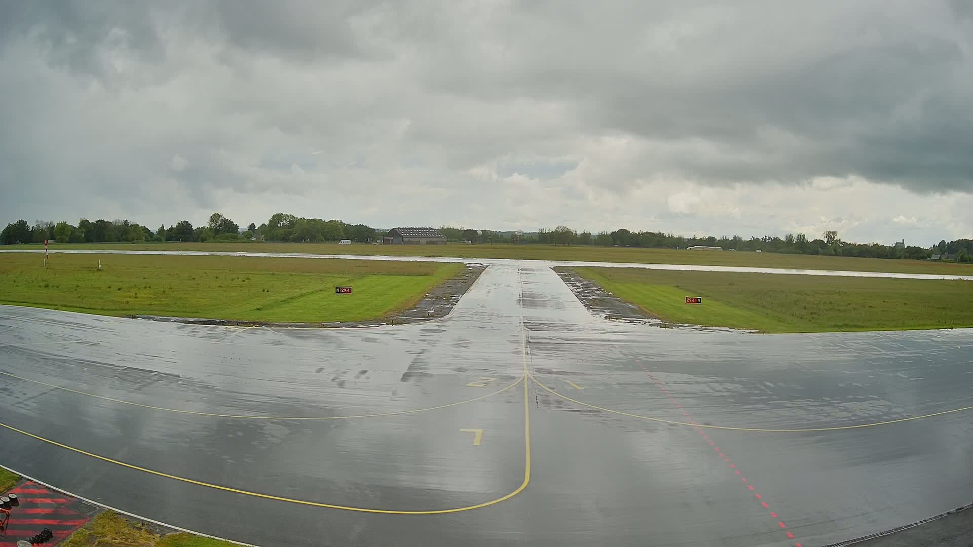 A wet, paved runway at a small airport stretches toward a grassy field under a cloudy sky.