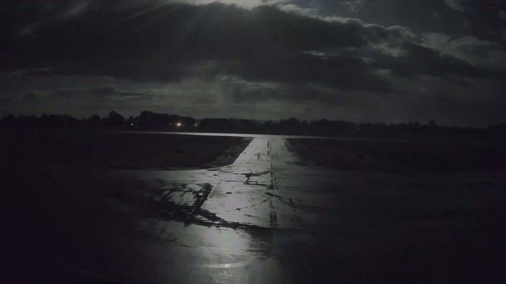 A wet, dark tarmac runway extends toward a moonlit, cloudy night sky.