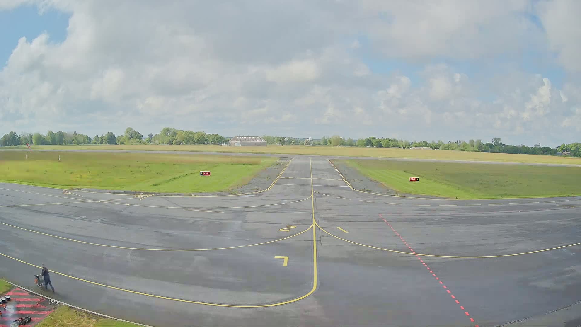 A wet airport runway and taxiway extend towards a grassy field under a partly cloudy sky.