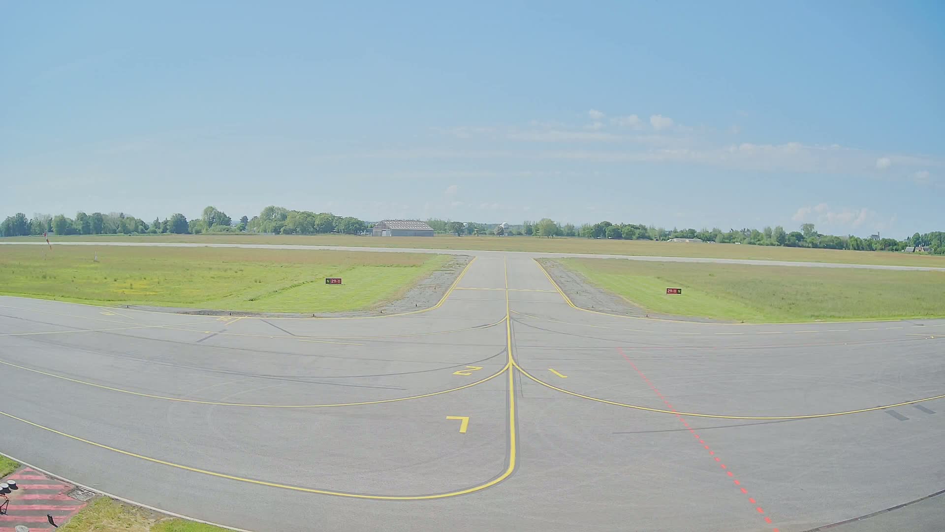 A paved runway at a small airport on a mostly sunny day extends into a grassy field bordered by trees.