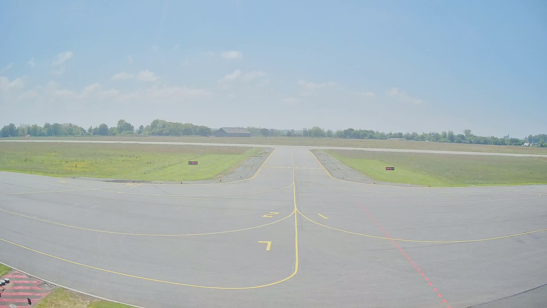A mostly empty airport tarmac on a partly cloudy day stretches toward a runway and a line of trees in the distance.