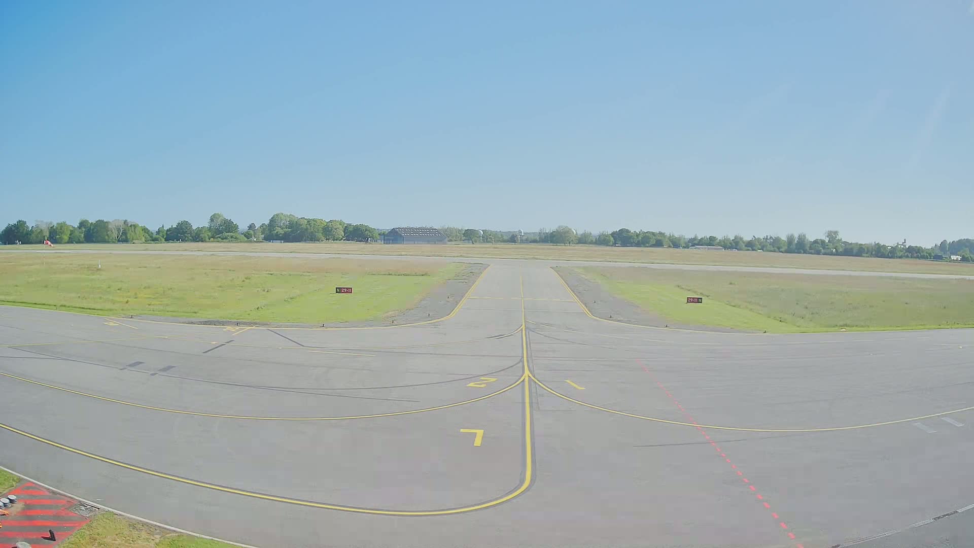 A tarmac apron at an airport on a sunny day, showing taxiway markings leading to a grassy runway and a line of trees in the distance.