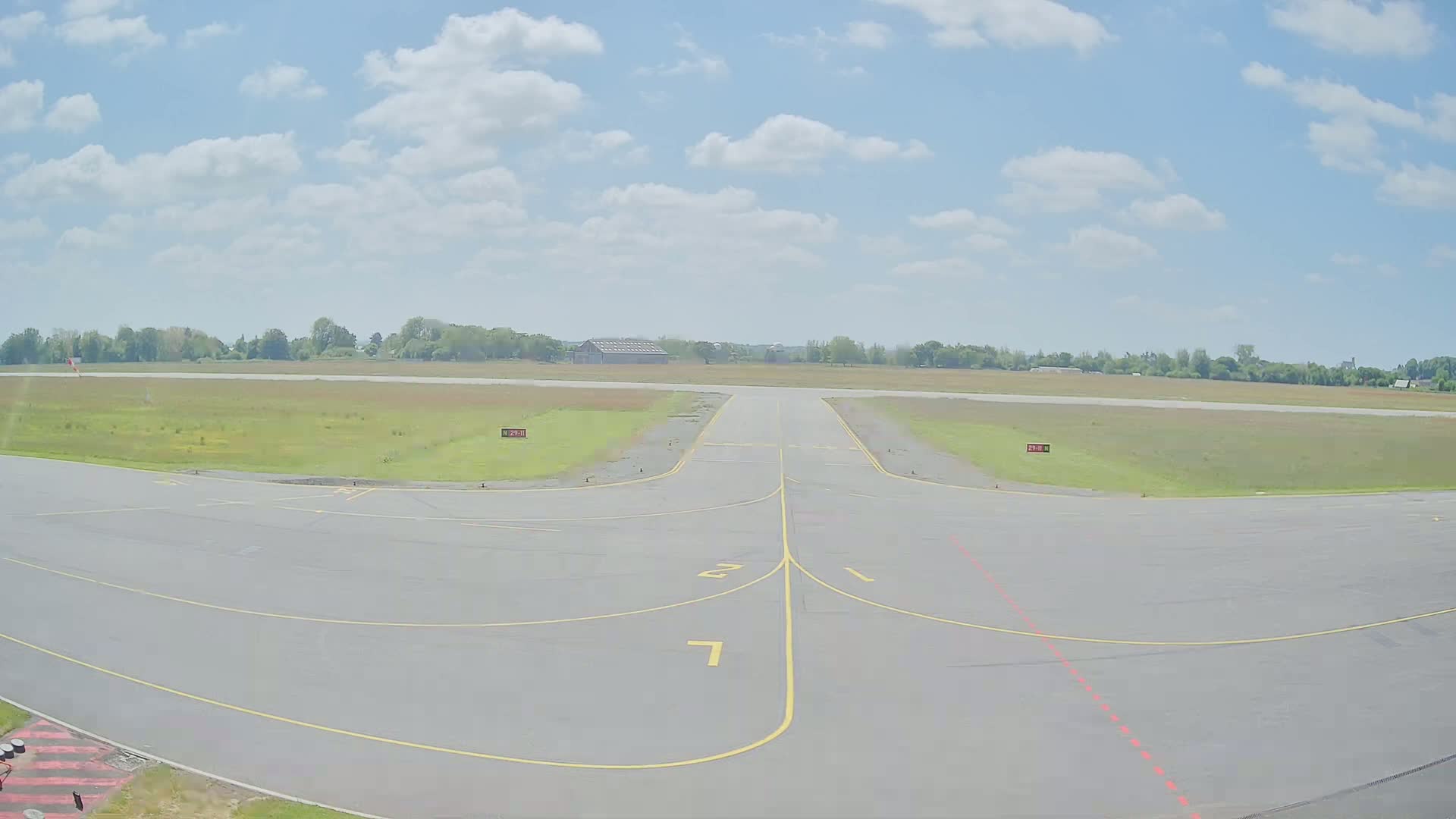 A tarmac area at an airport on a partly cloudy day shows a runway and taxiways leading to a grassy field and trees in the distance.
