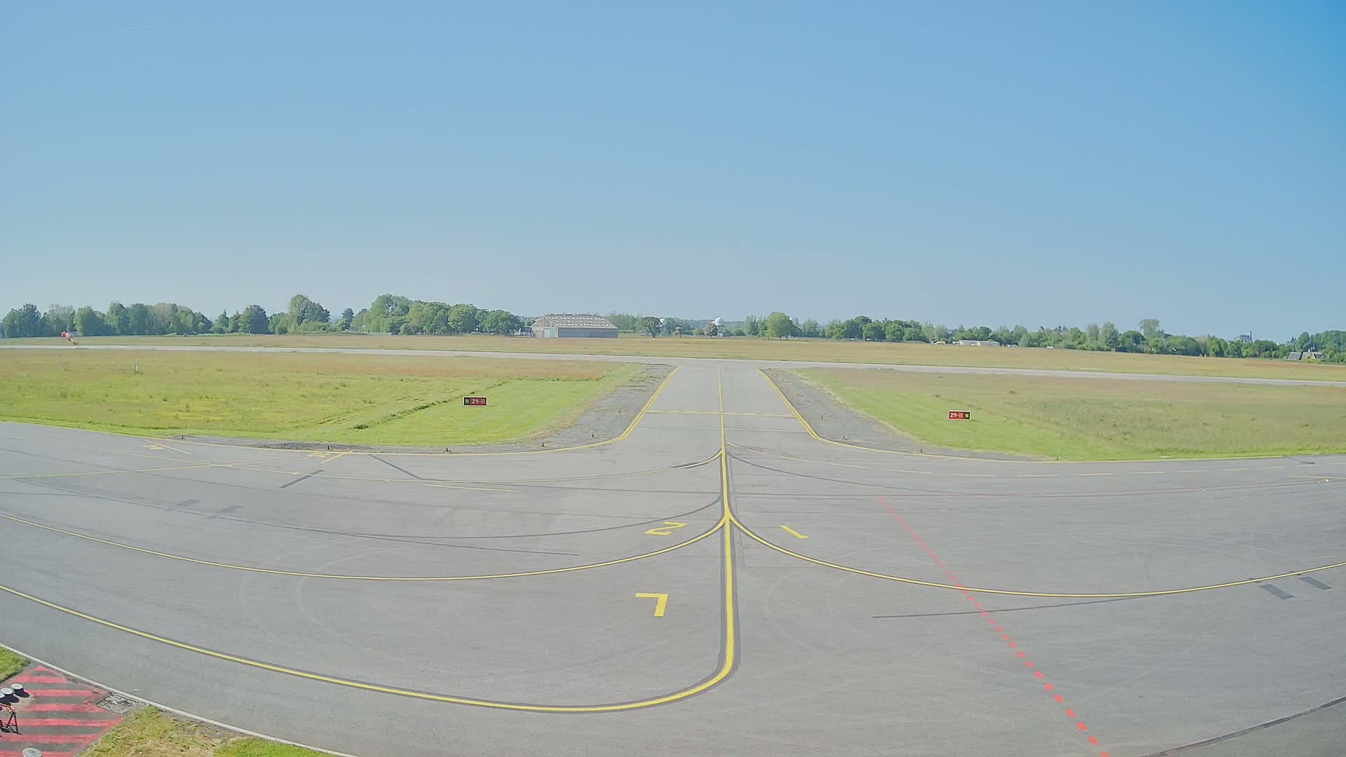 A tarmac area of an airport on a clear, sunny day, with a runway extending to a grassy field bordered by trees in the distance.