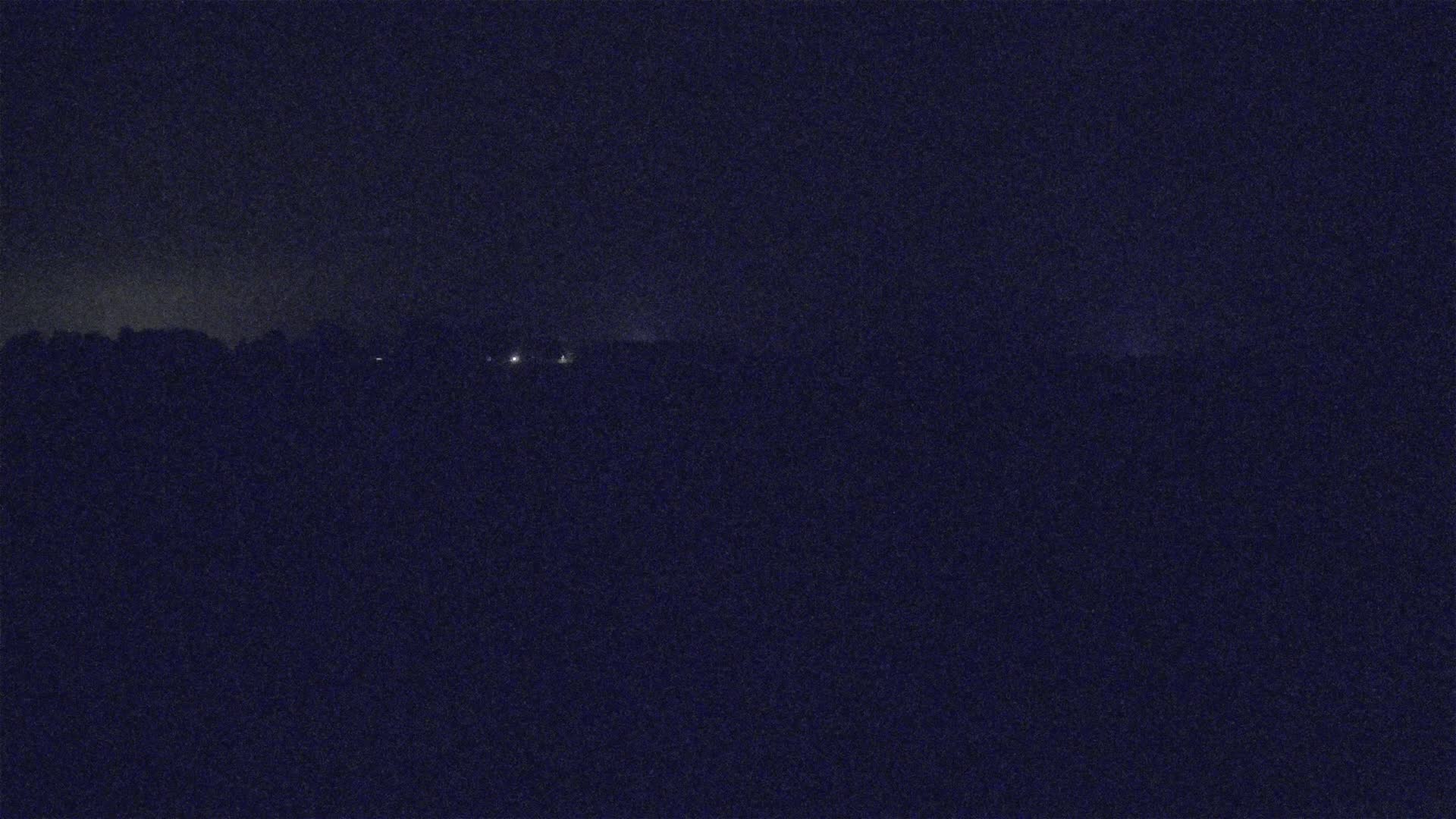 A dark, nighttime landscape shows a line of distant lights behind a dark, silhouetted treeline under a cloudy sky.