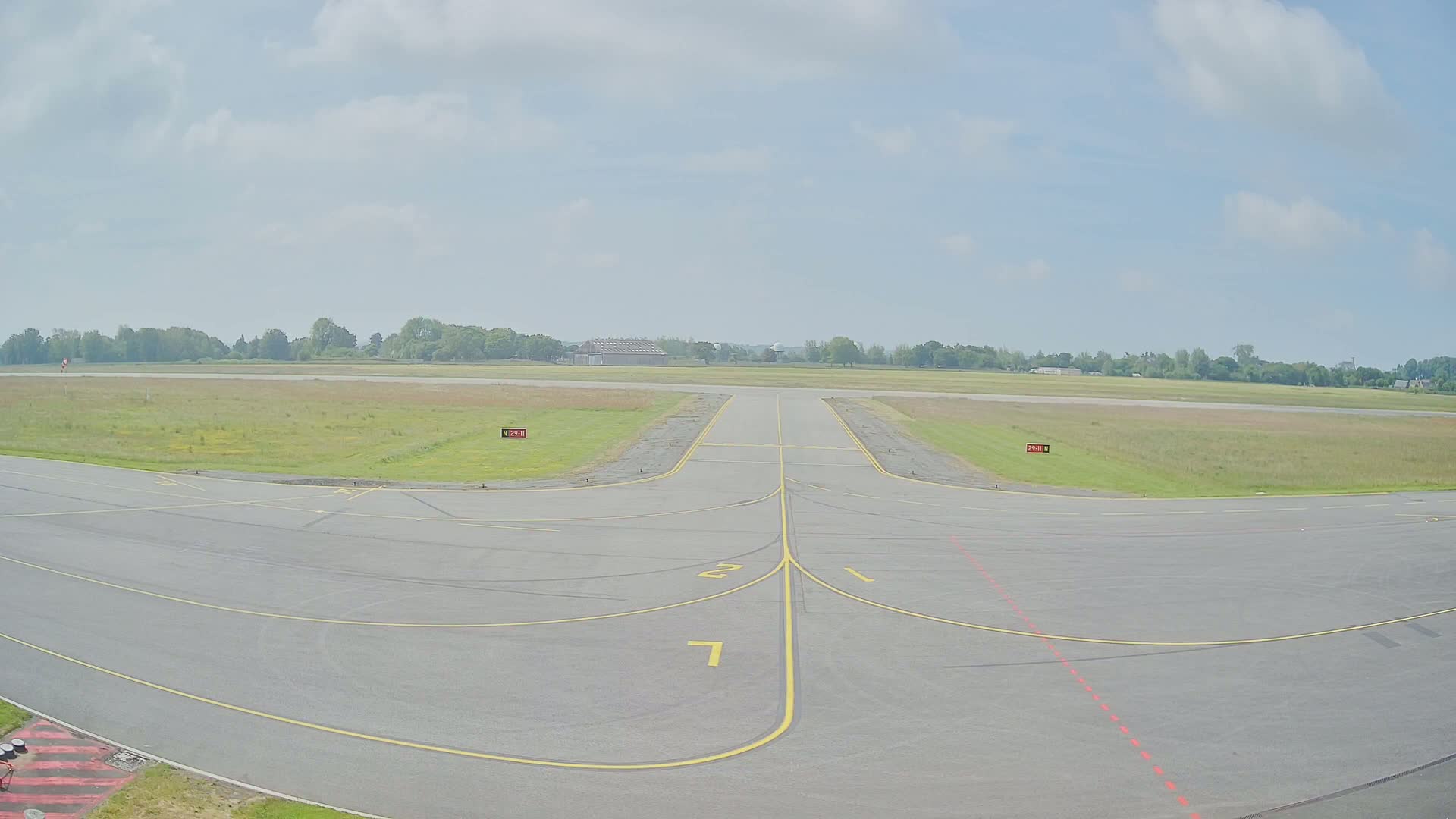 A paved airport tarmac with yellow lines marking taxiways extends toward a grassy airfield under a partly cloudy sky.