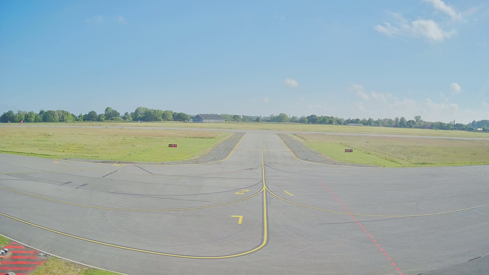 A paved airport runway with yellow lines and a grassy field under a mostly clear blue sky.