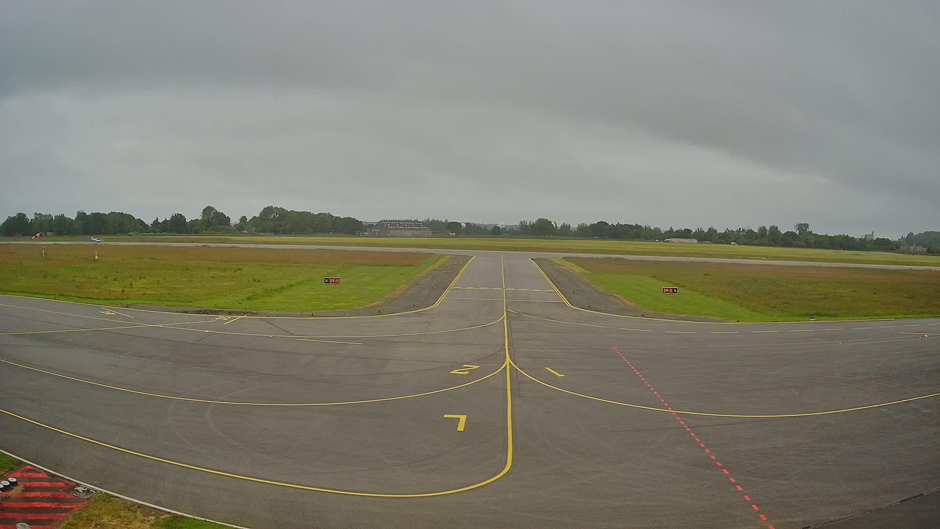 An overcast sky hangs over an airport runway, where the tarmac intersects with grassy fields and a small airplane is visible in the distance.