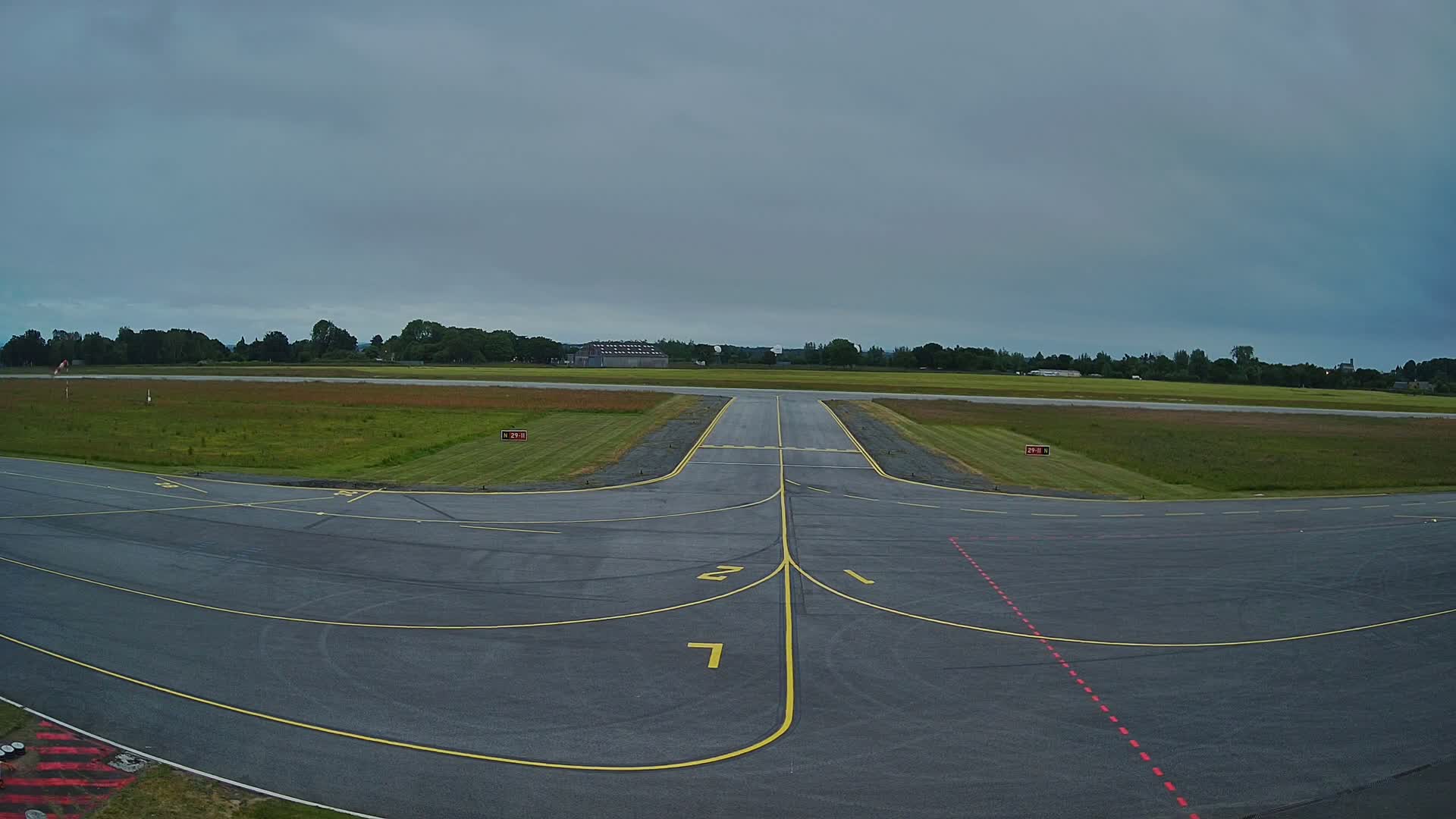 A gray tarmac airport runway with yellow lines under an overcast sky, surrounded by green grass and trees.