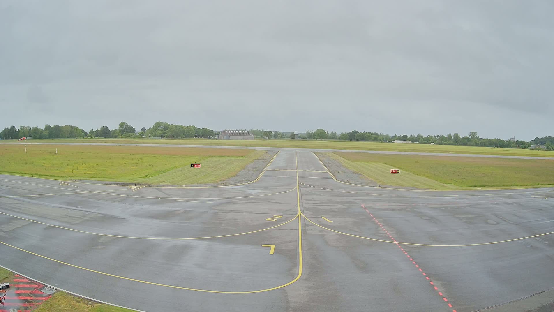A wet airport runway extends toward a grassy field under a cloudy sky.