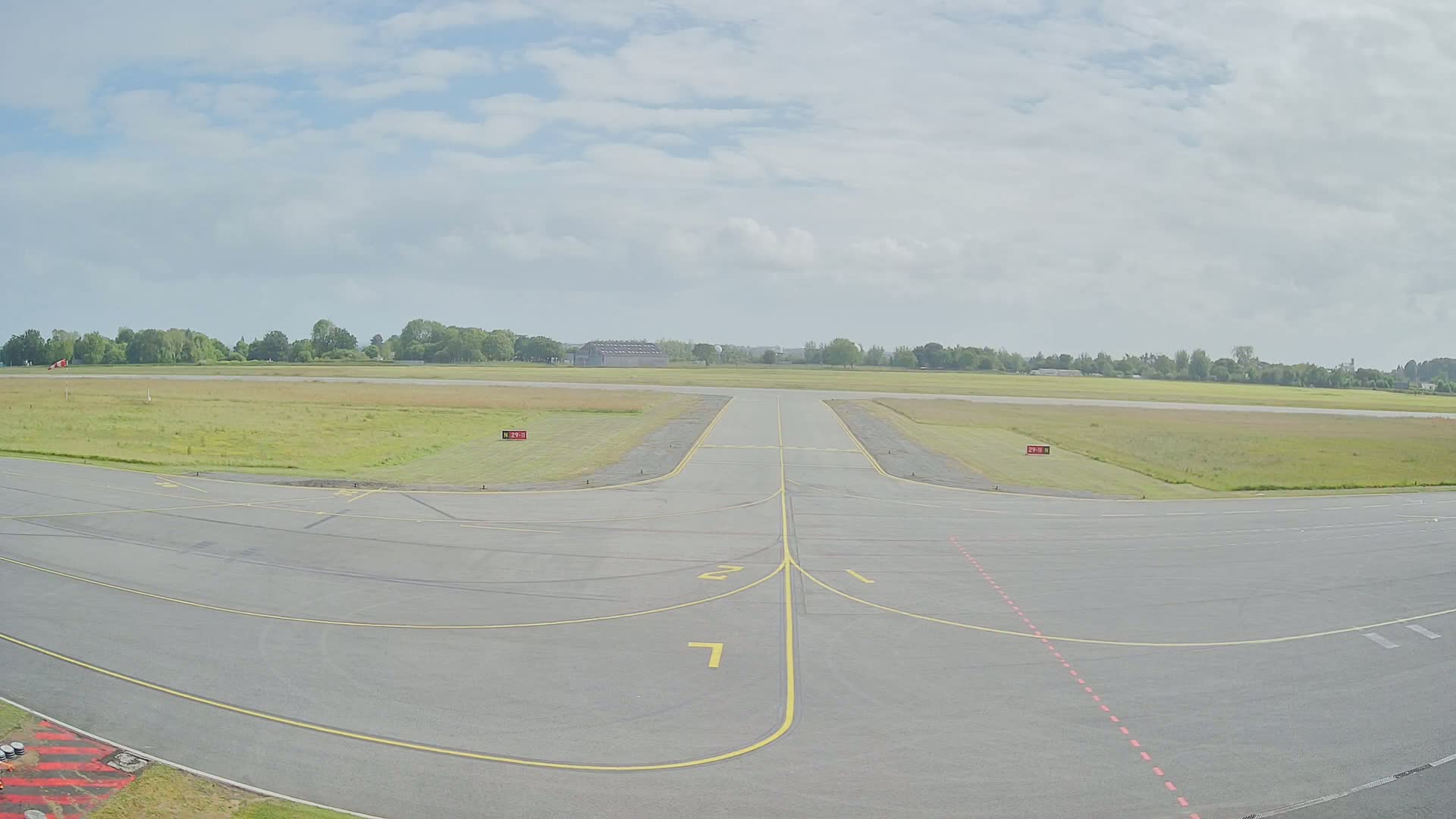 A paved airport runway and taxiway extend toward a grassy field under a partly cloudy sky.