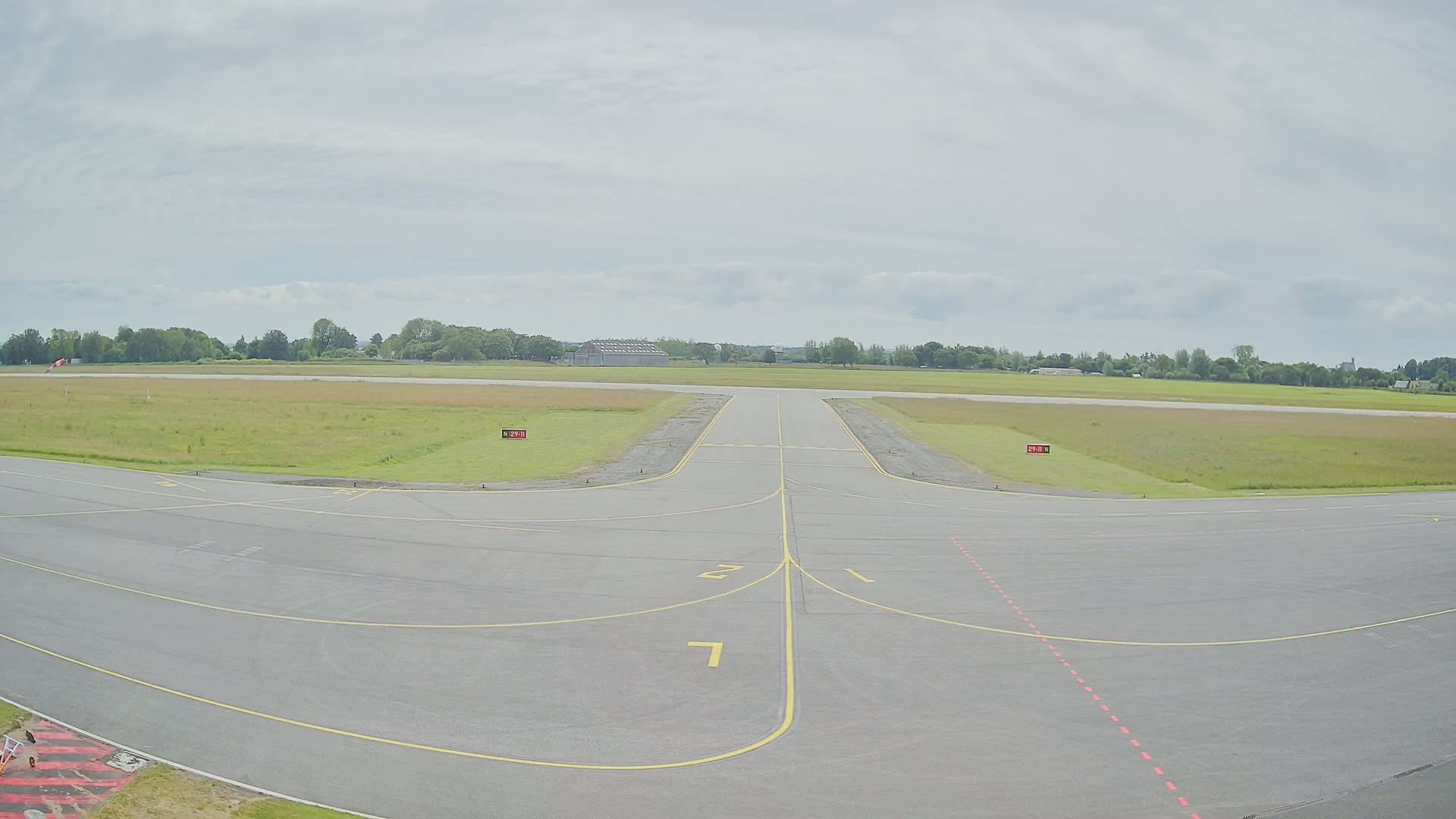 A paved airport runway with yellow and red markings stretches toward a grassy field under a cloudy sky.