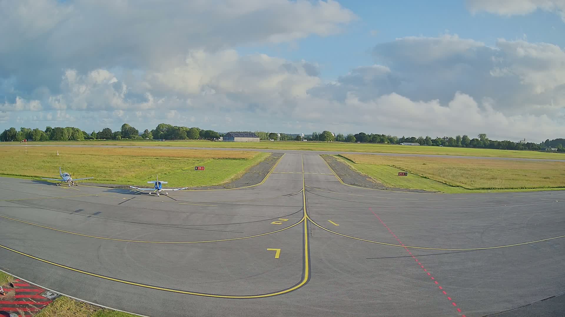 Two small airplanes are parked on a tarmac at a small airport under a partly cloudy sky.
