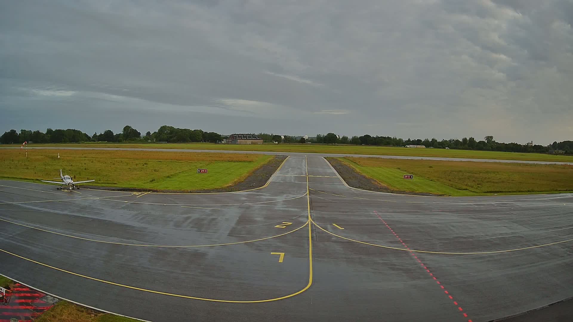 A small airplane sits on a wet tarmac runway at a small airport under a cloudy sky.
