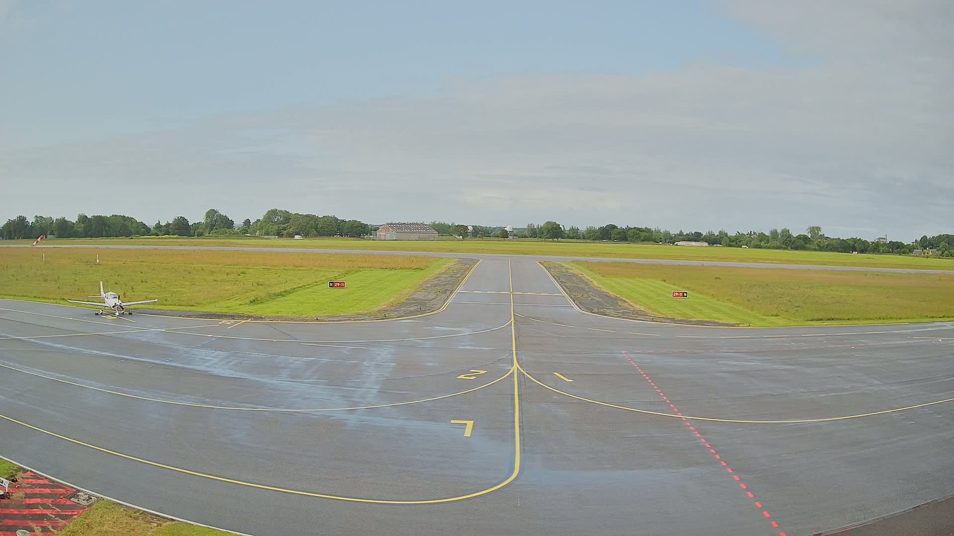 A small airplane sits on a wet tarmac at the intersection of airport runways, under a mostly cloudy sky.