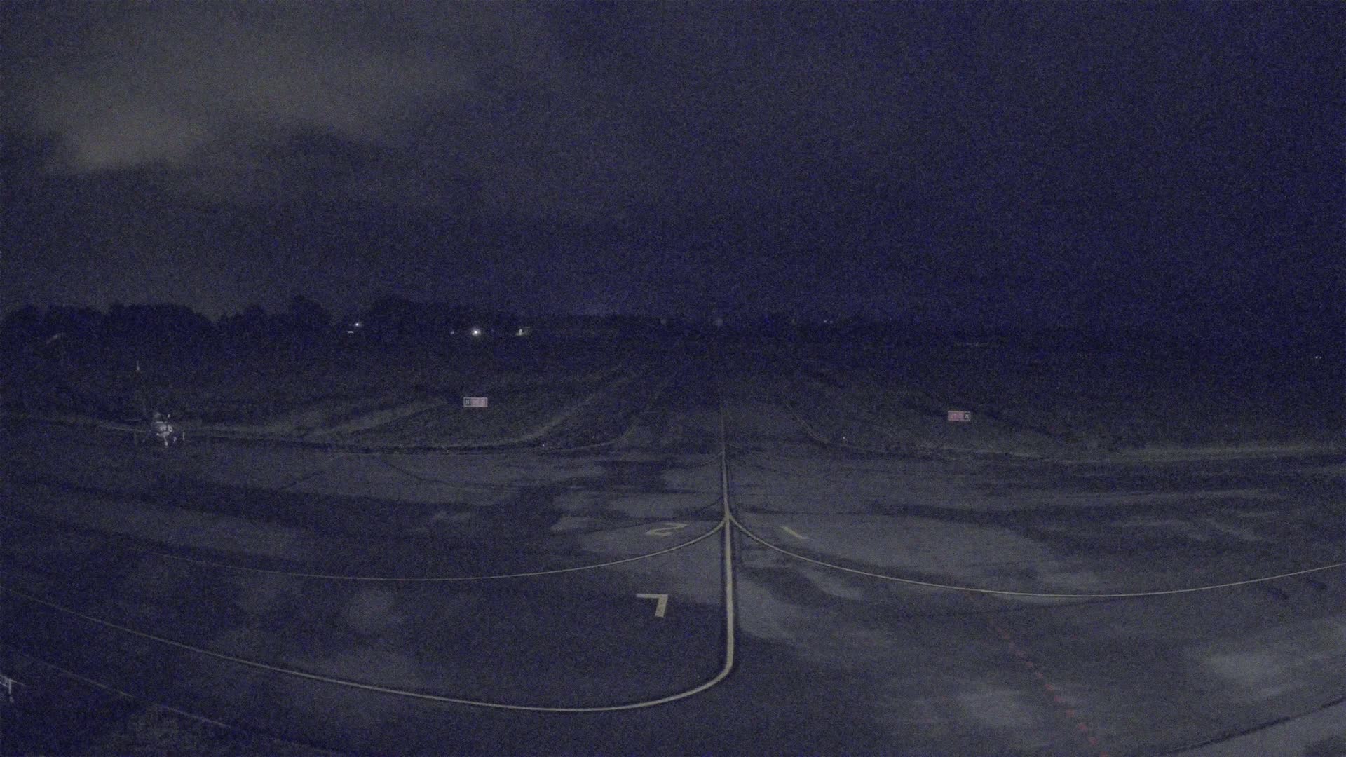 A dark, nighttime view of a runway with faint yellow markings, under a cloudy sky.