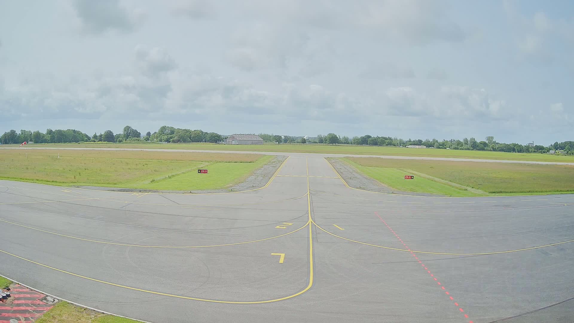 A tarmac runway at an airfield under a partly cloudy sky, leading to a grassy field and trees in the distance.