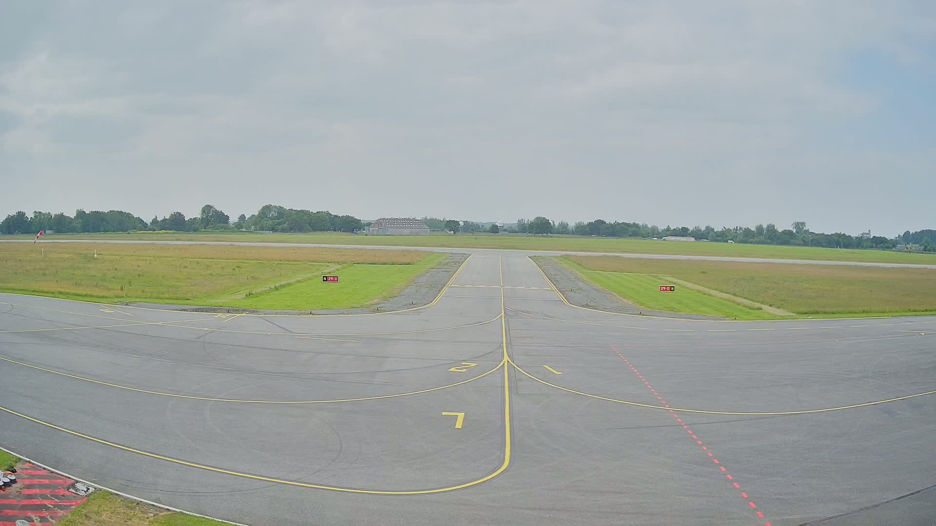 A tarmac runway at an airfield under a cloudy sky, with grassy fields extending to a line of trees in the distance.