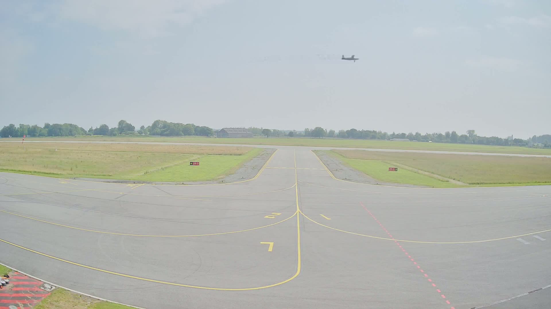 A small airplane flies over a tarmac at an airfield on a partly cloudy day.