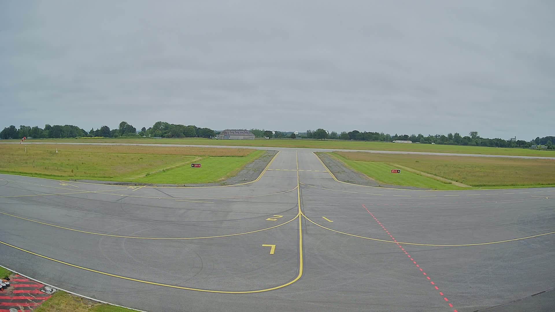 An airport tarmac under an overcast sky shows a runway and taxiway markings leading to a grassy field bordered by trees.