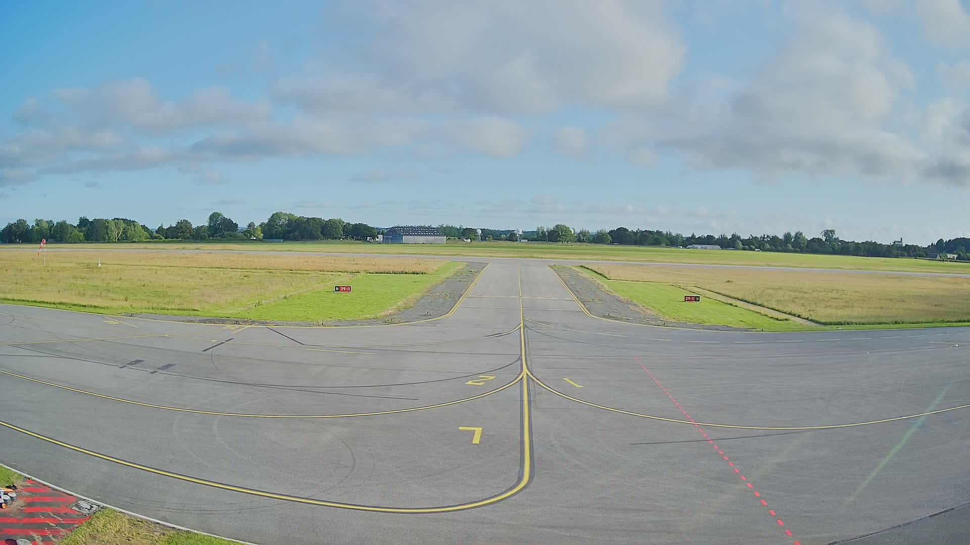 A paved airport runway and taxiway area under a partly cloudy sky extend to a grassy airfield, bordered by trees in the distance.