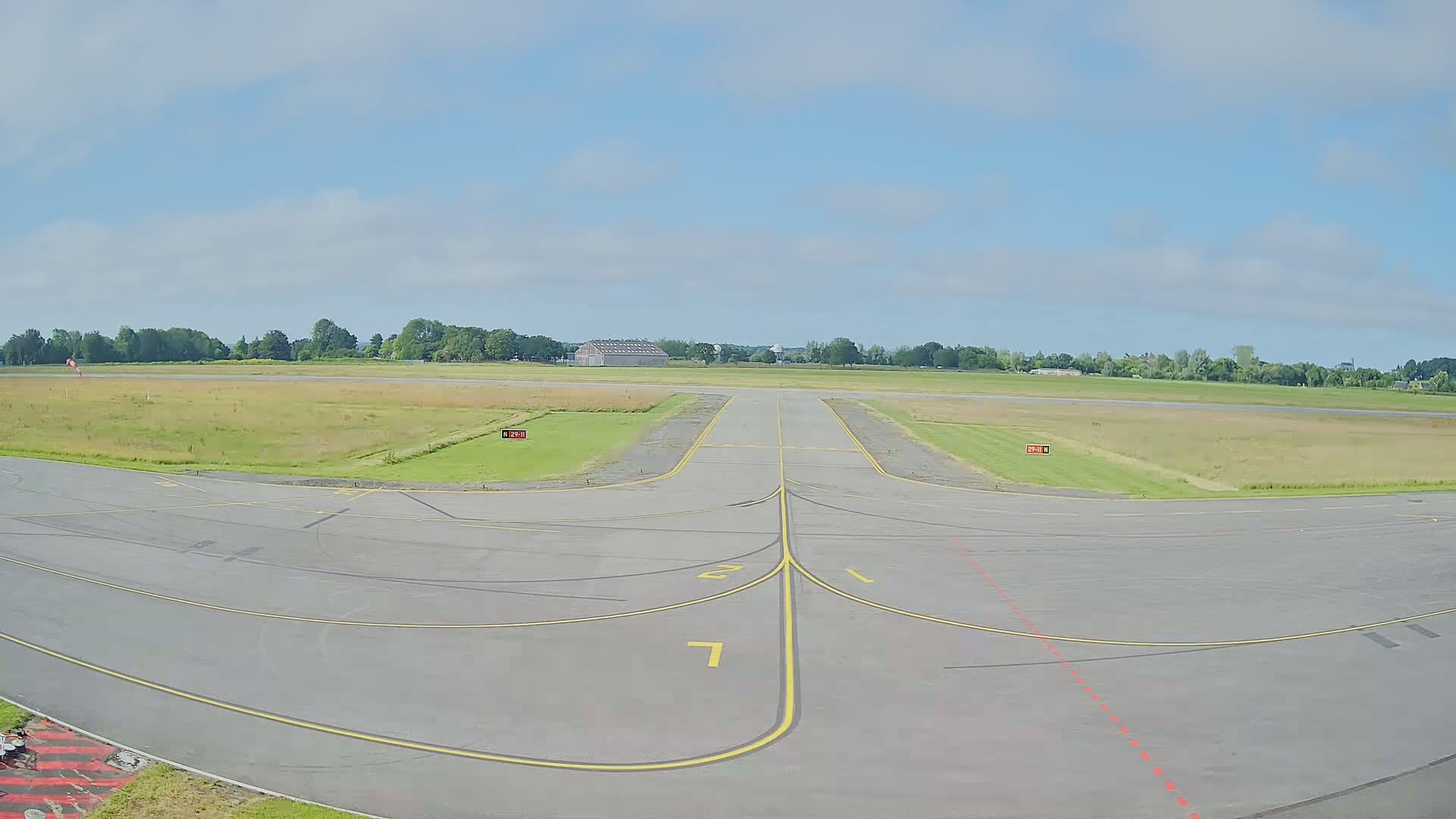 A paved airport runway and taxiway area extends to a grassy airfield under a partly cloudy sky.