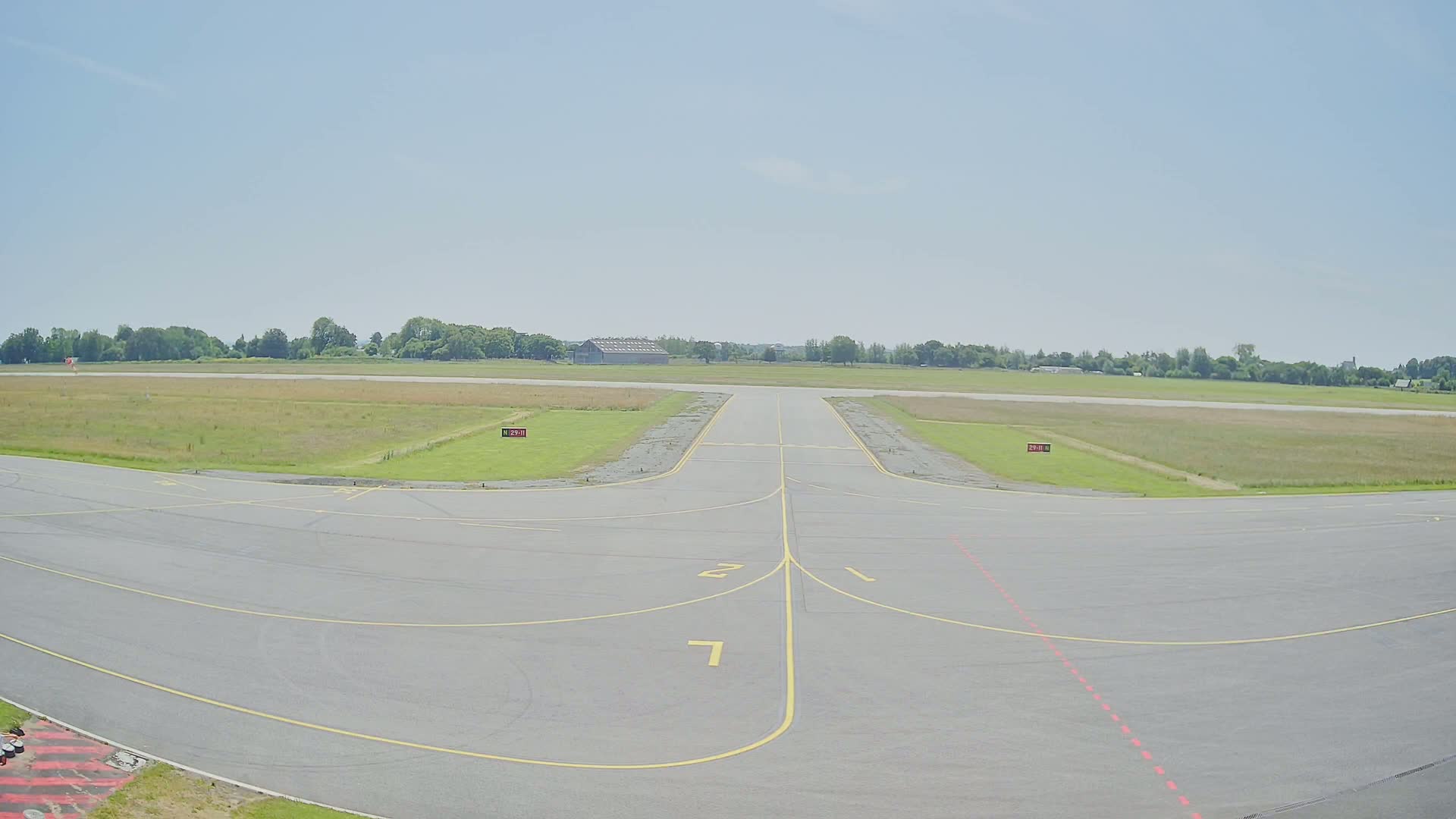 A paved airport runway with yellow lines extends toward a grassy field under a clear, bright sky.