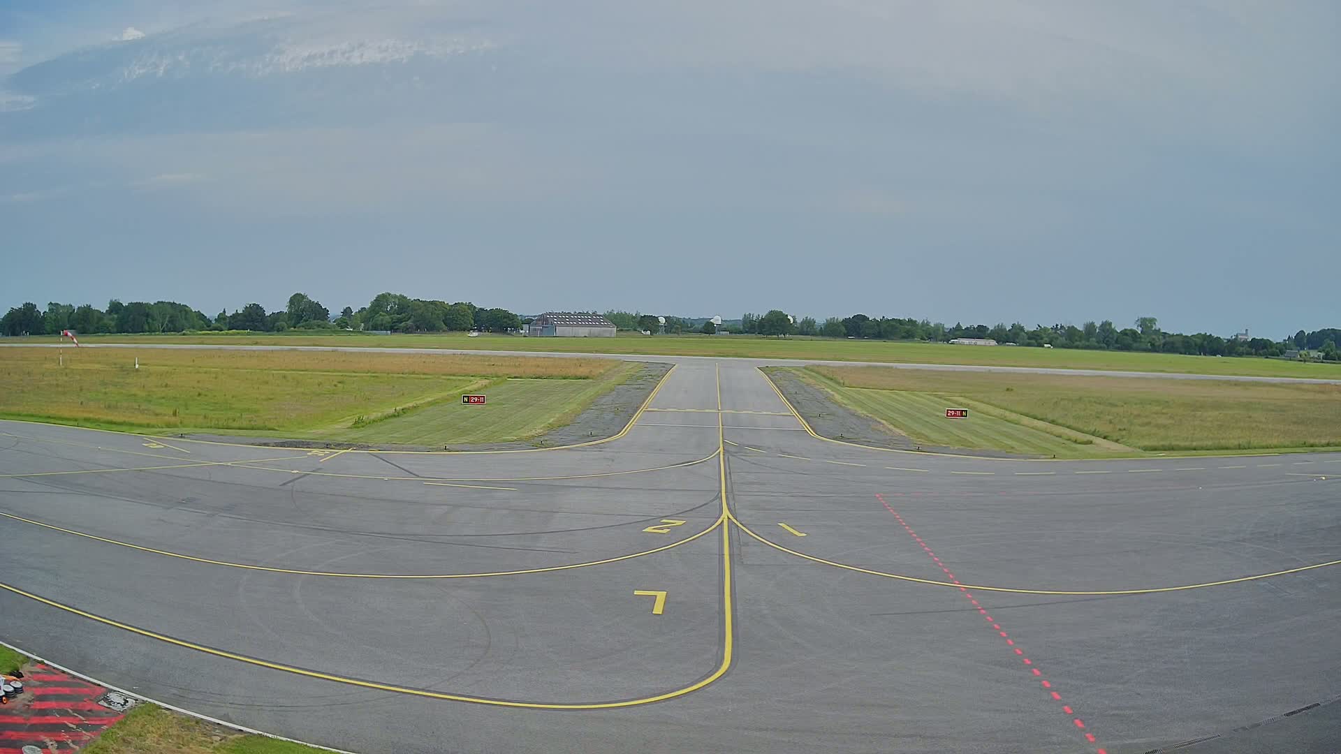 An airport runway with short grass and trees in the background under a cloudy sky.