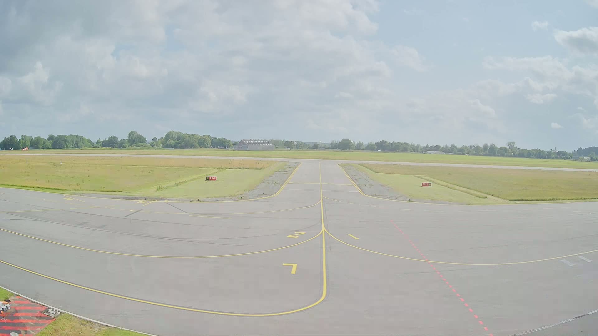 A tarmac runway with yellow lines curves toward a grassy airfield under a partly cloudy sky.