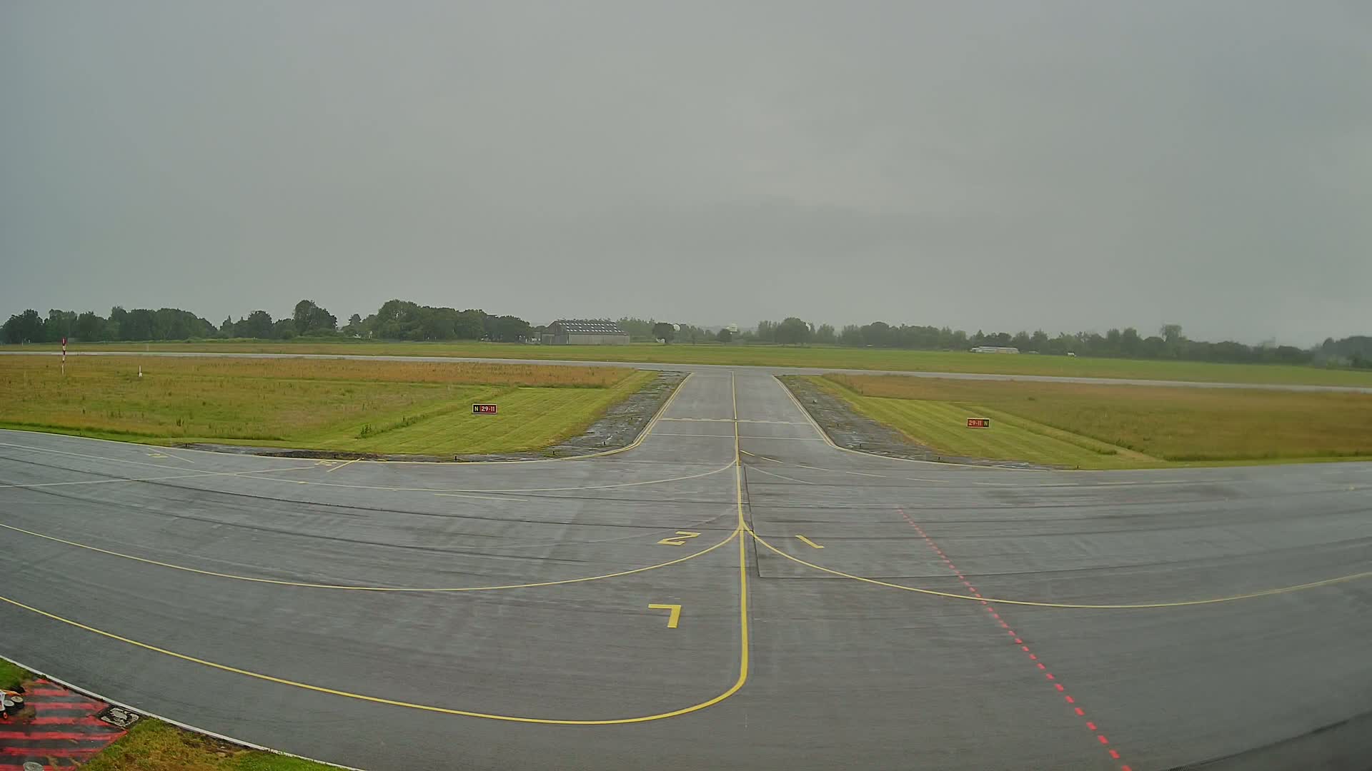 A wet airfield runway with yellow lines stretches into the distance under an overcast sky.