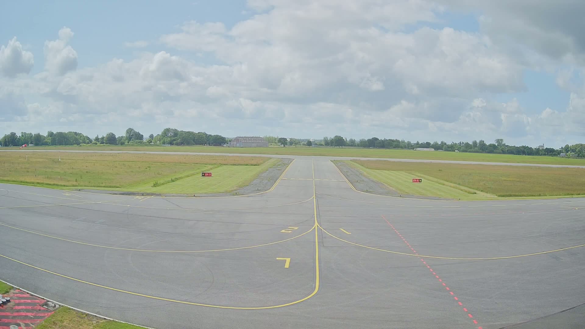 A mostly sunny day at an airport runway shows a paved tarmac with yellow lines leading to a grassy field beyond.