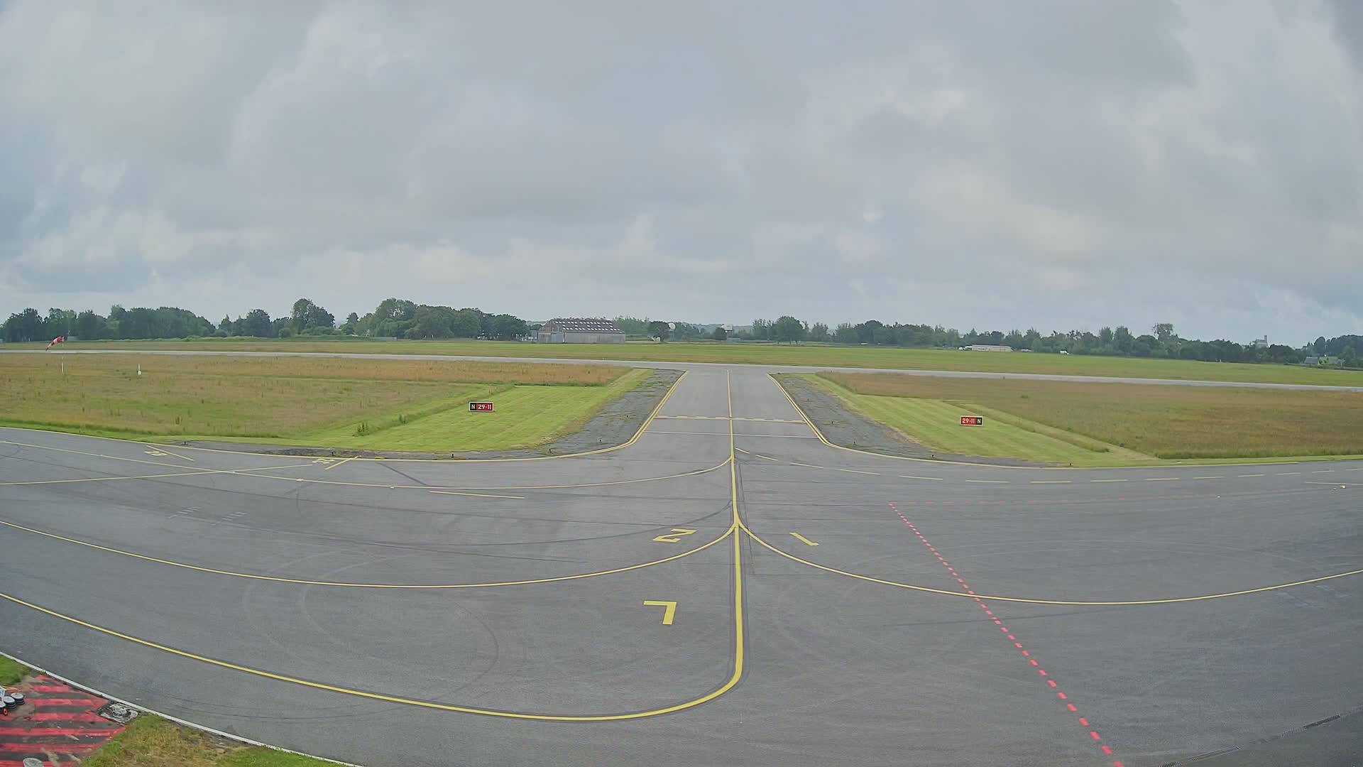 An overcast sky hangs over a tarmac area with yellow lines marking a runway and taxiway, leading to a grassy field bordered by trees.