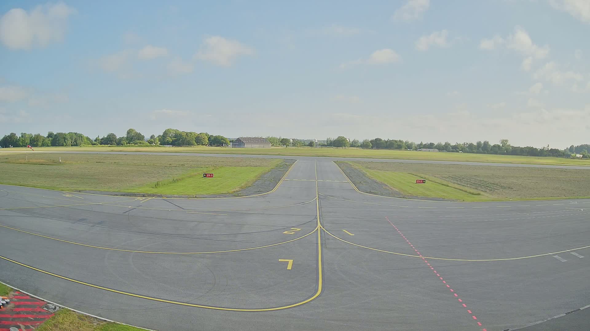 A mostly sunny day shows a tarmac area with runway markings at a small airfield, surrounded by grassy fields and a line of trees in the distance.