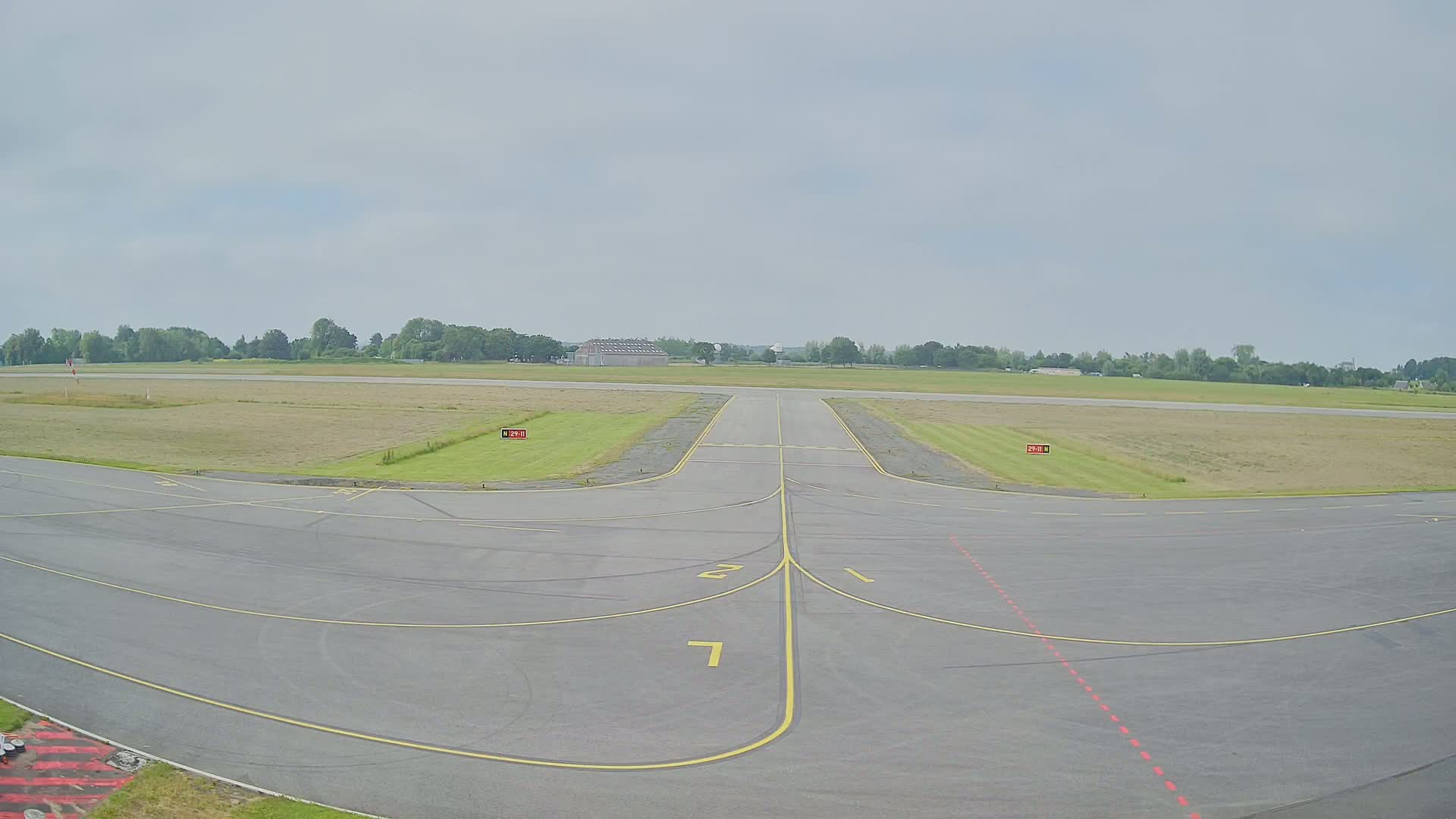 An empty airport tarmac under an overcast sky extends to a runway leading to a grassy field and trees in the distance.