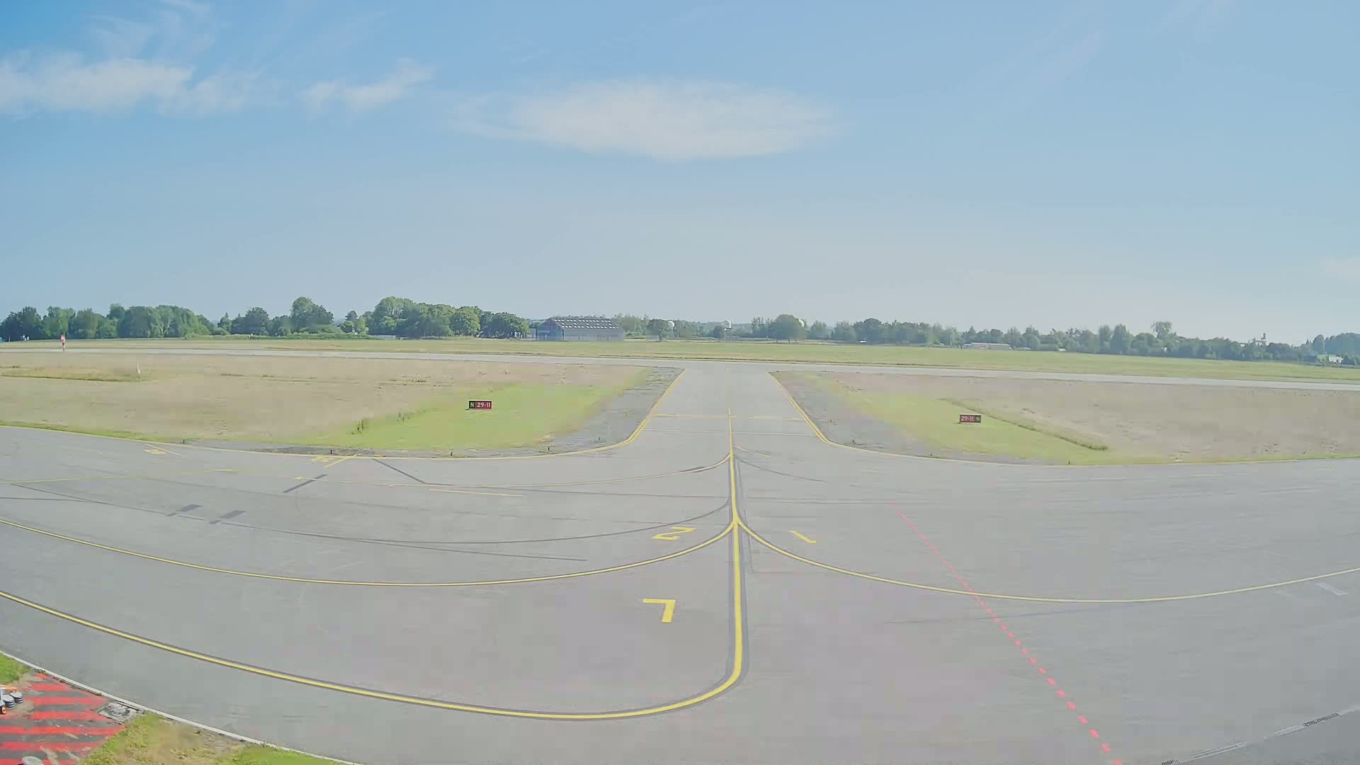 A paved airport taxiway with yellow lines curves toward a grassy runway under a mostly clear, bright sky.
