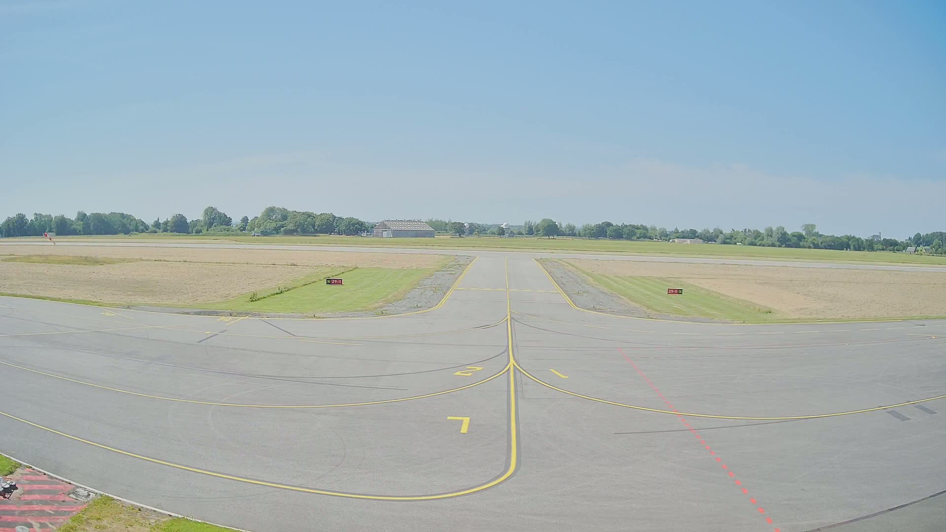 An airport runway with yellow lines marking taxiways extends into a field under a clear, sunny sky.