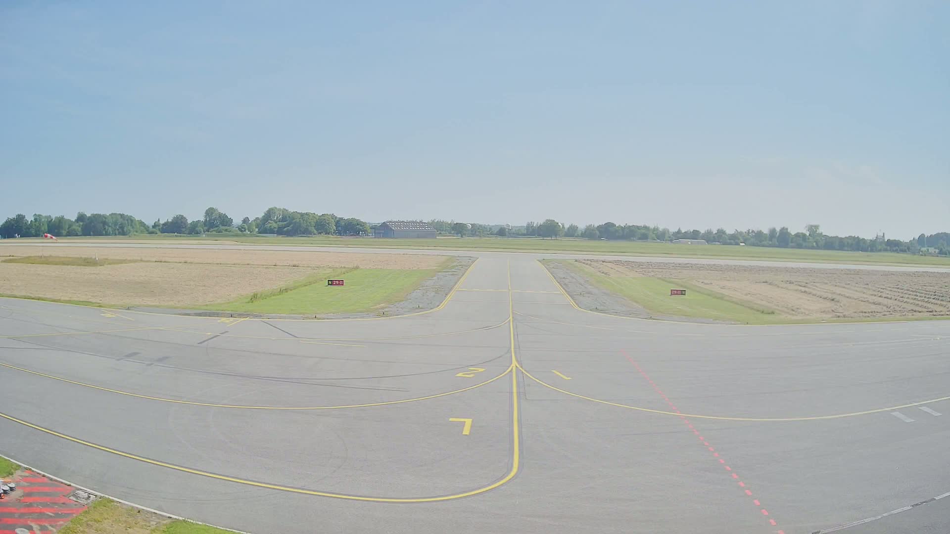 A tarmac airport runway and taxiway area stretches toward a treeline under a clear, sunny sky.