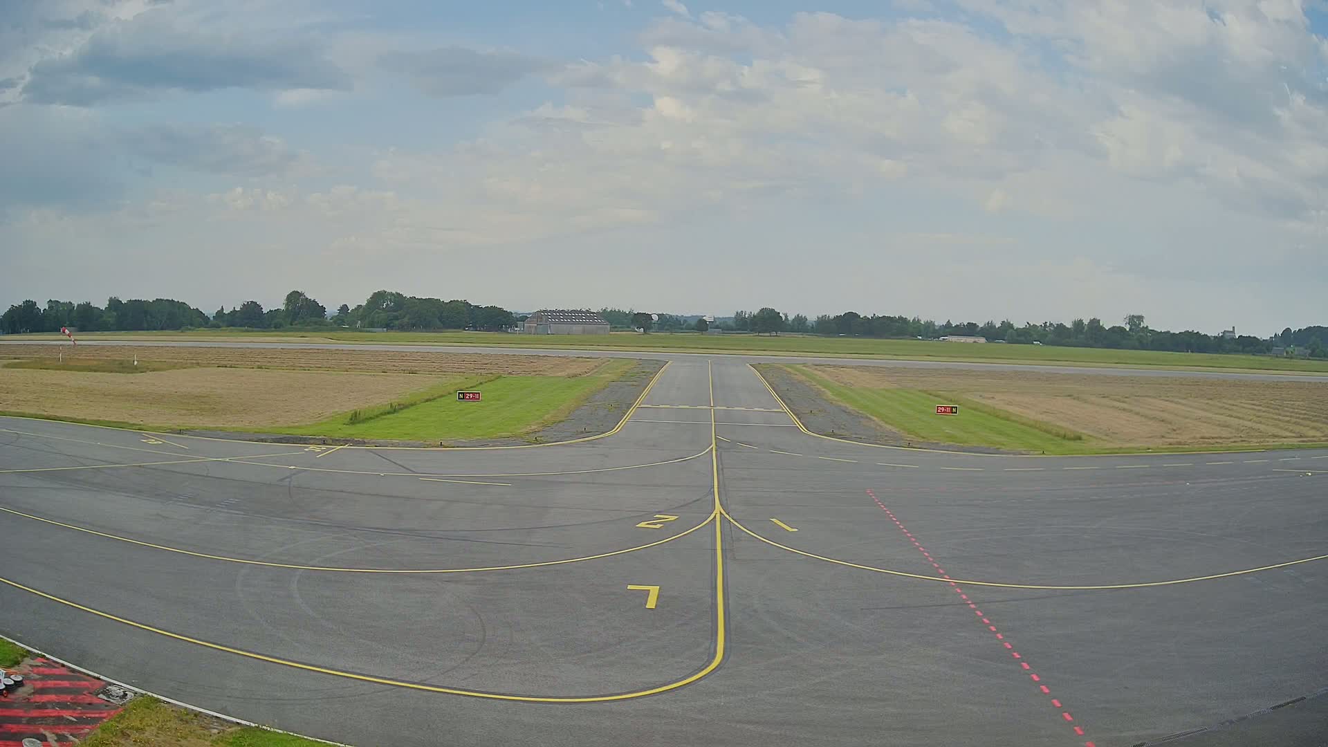 An airport tarmac under a partly cloudy sky shows a runway leading to a grassy field bordered by trees.