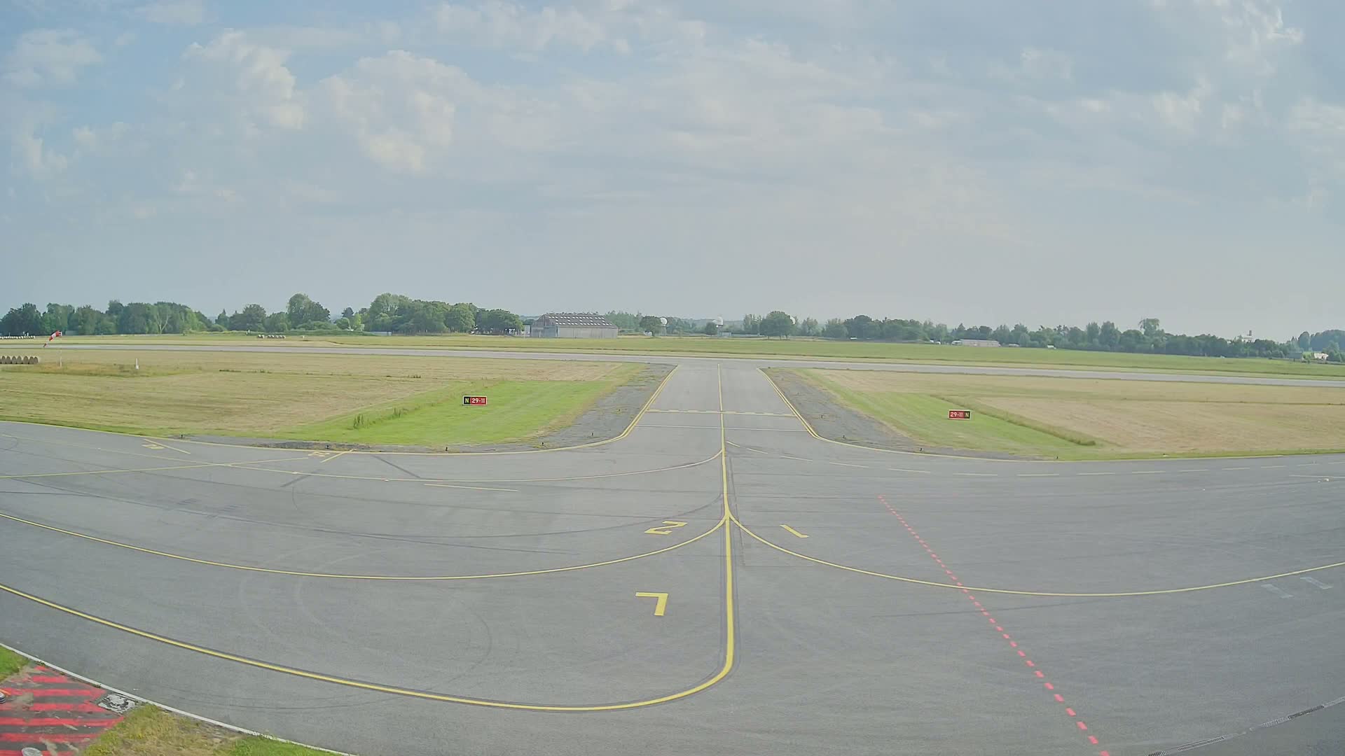 A paved airport taxiway under a partly cloudy sky leads to a grassy runway surrounded by trees.