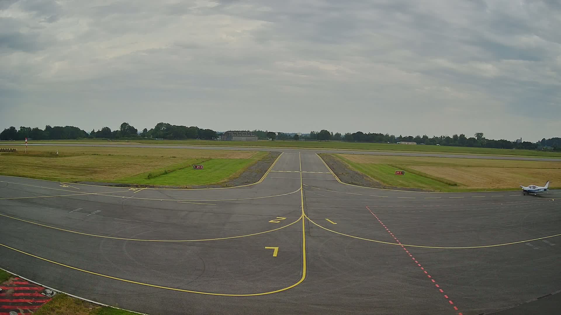A small airplane sits on an airport tarmac under an overcast sky, with a runway extending toward a line of trees in the distance.