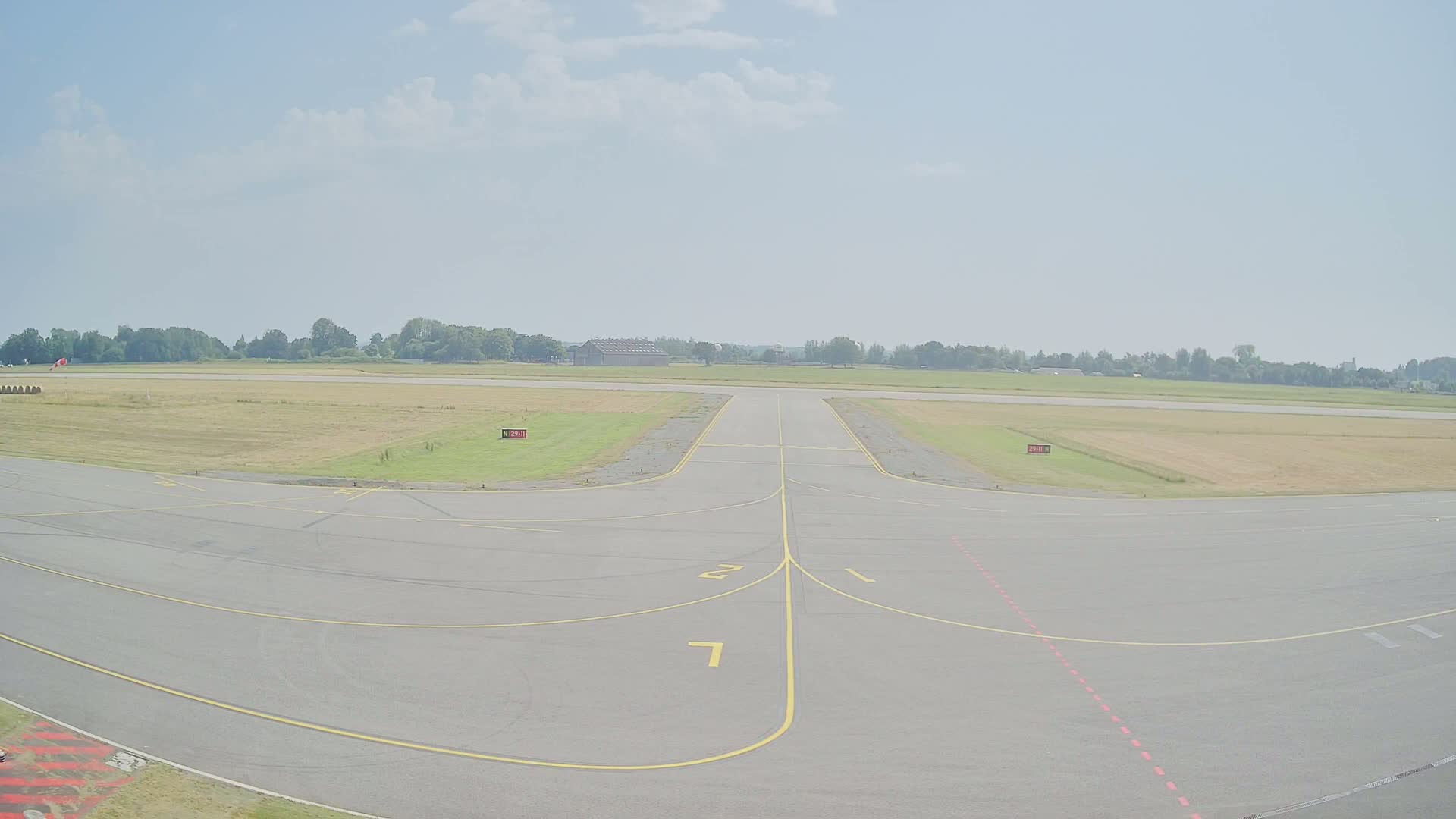A paved airport runway with yellow lines extends toward a grassy field under a mostly clear sky.
